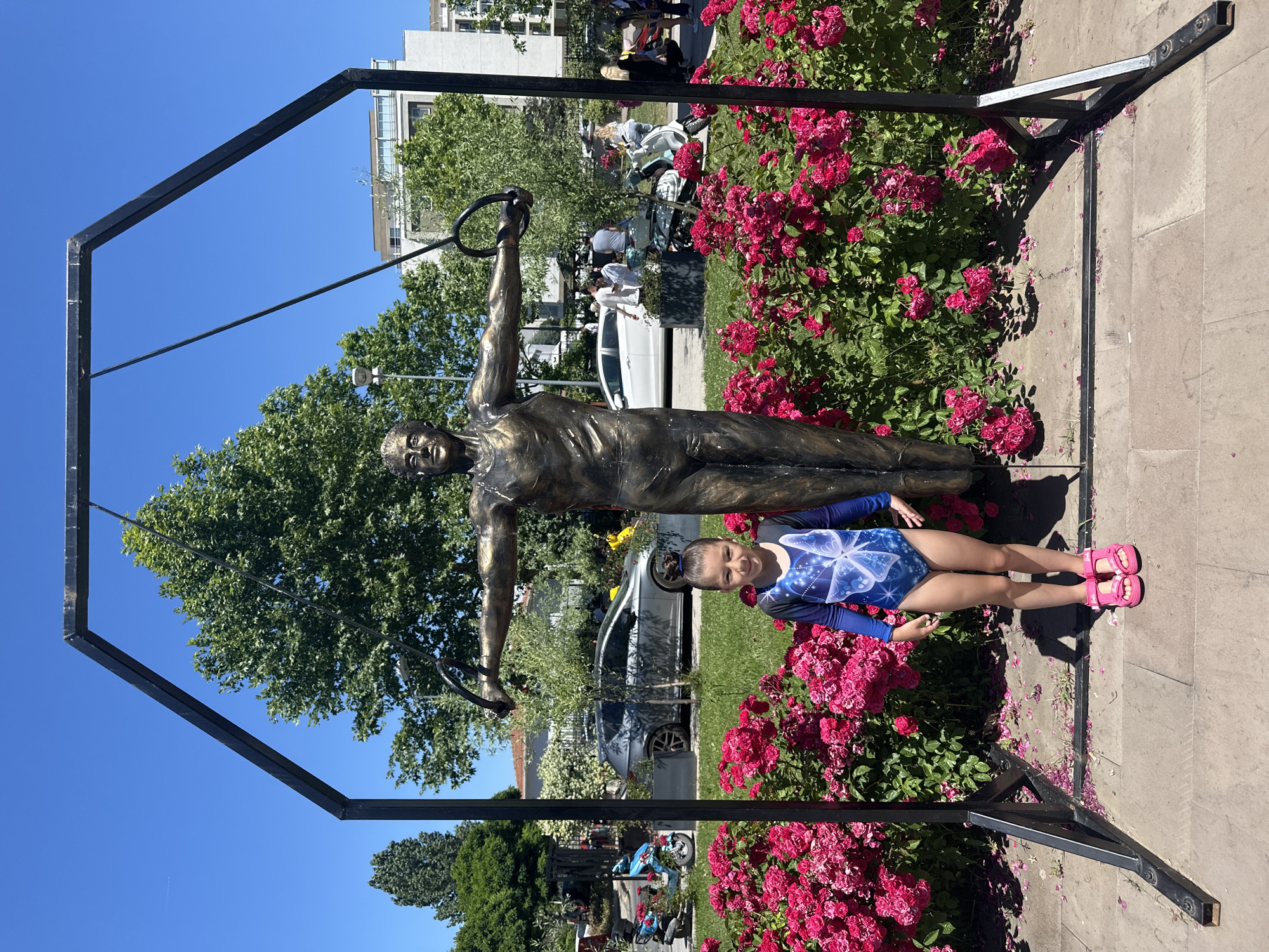 The image features a young girl posing next to a bronze statue of a gymnast holding rings. The statue is positioned in front of a vibrant bed of pink roses. The girl, wearing a blue and white leotard with a butterfly design and pink sandals, is smiling at the camera. Behind the statue and the girl, a street scene is visible, with cars and trees. In the background, modern buildings can be seen under a clear blue sky. The overall atmosphere suggests a sunny day, likely during the daytime, in a public outdoor space like a park or square. The setting, identified as Sancaktepe, Turkey, implies a specific geographical and cultural context. The image captures a moment of playful interaction between a child and a public monument, set against a backdrop of urban life and nature.