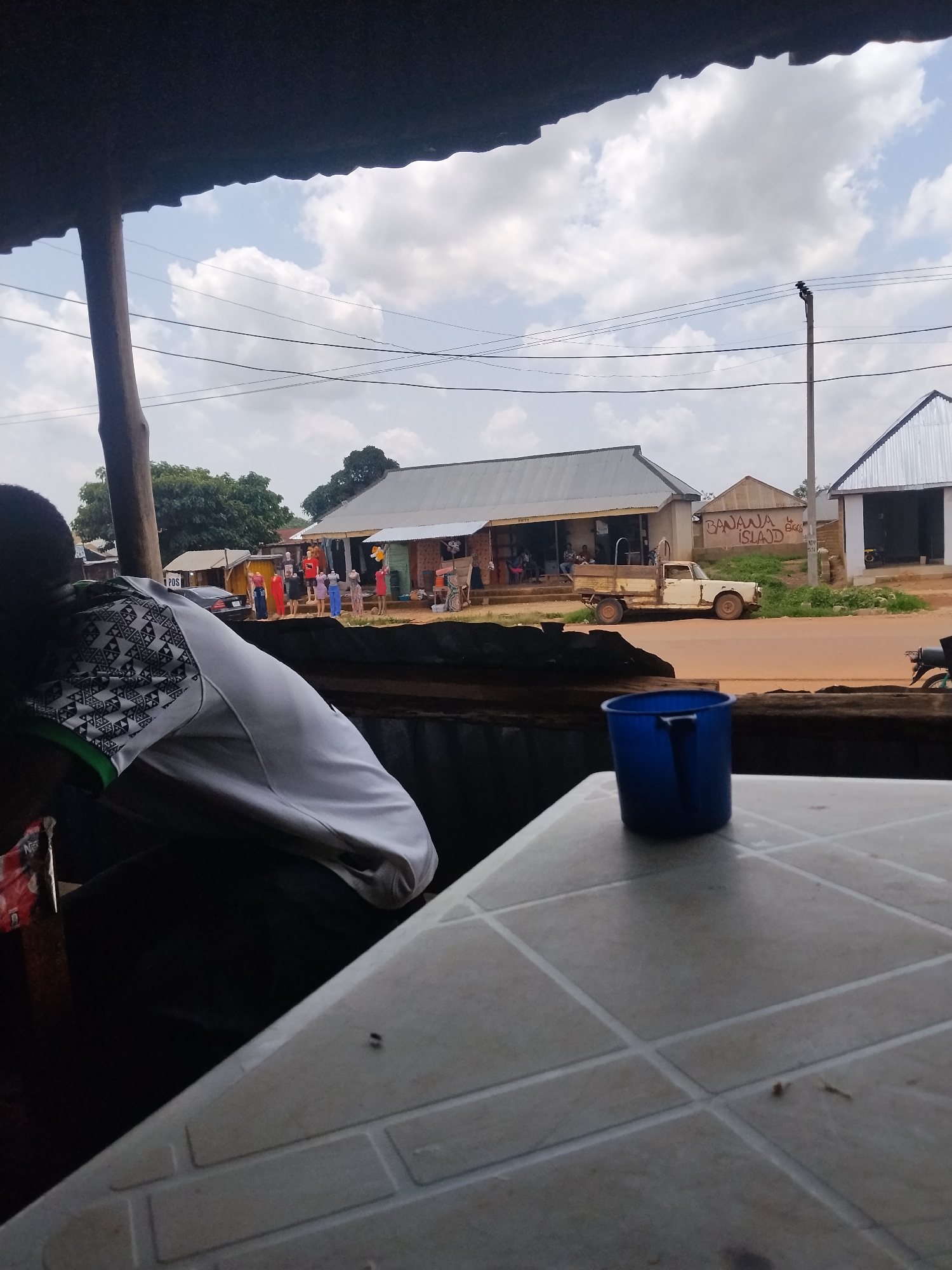 The image depicts an outdoor scene, likely a roadside stall or small market, in Durumi, Nigeria. In the foreground, a person wearing a white shirt with a black geometric pattern is partially visible, seated on the left. In front of them is a table with a tiled surface, on which rests a blue plastic cup. The background reveals several buildings with corrugated metal roofs, suggesting a commercial area. A light-colored pickup truck is parked on a dirt road in front of one of the buildings. Several mannequins dressed in colorful clothing are displayed outside another building, indicating a clothing shop. A utility pole with wires runs across the upper part of the image, and trees are visible in the distance. The sky is partly cloudy, suggesting daytime. The overall impression is of a typical street scene in a Nigerian town.