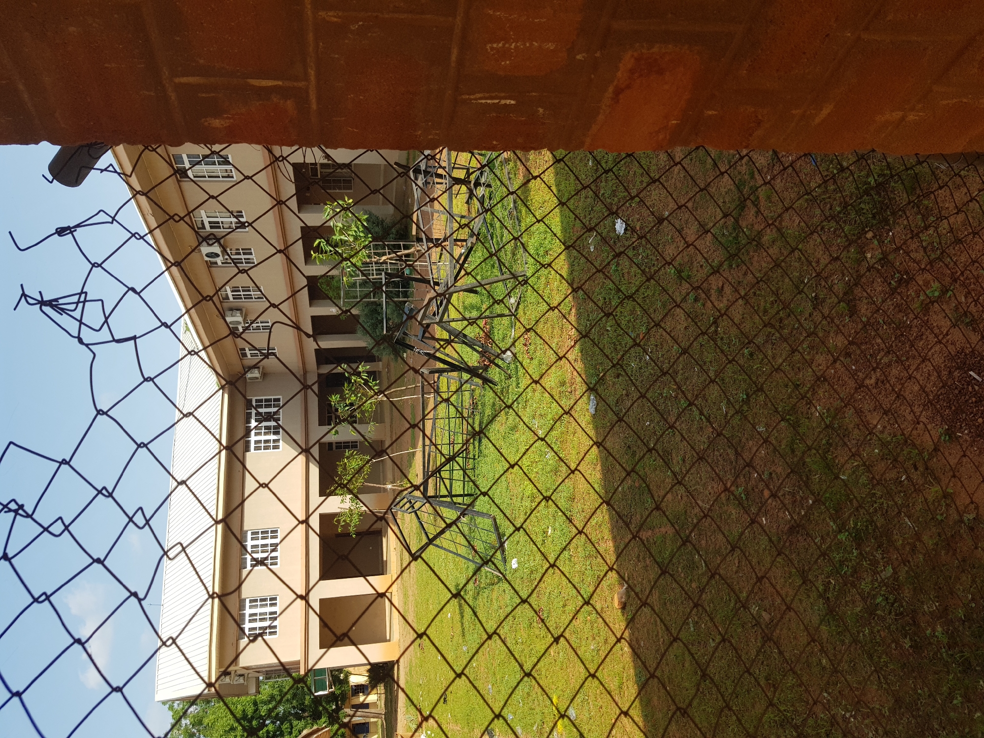 A wire fence with a rusty brick pillar on the right obscures a view of a courtyard with a two-story building in the background. The building is beige with white-framed windows, a white roof, and covered walkways on the ground floor. The courtyard is covered in green grass, with some scattered debris. In the middle of the courtyard, several black metal pieces appear to be parts of broken playground equipment. A small, light-colored animal is lying on the grass in the lower left quadrant of the image. The sky is clear and blue. The scene is likely located in the city of Jimeta, Nigeria, given the architectural style and vegetation.