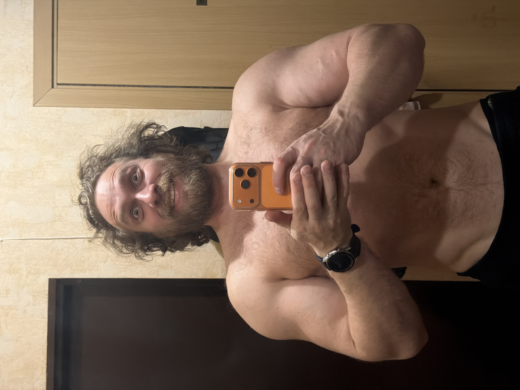 A man with long, curly brown hair and a full beard takes a mirror selfie in an indoor setting. He appears to be in his 30s or 40s. The man is shirtless, revealing his muscular upper body and chest hair. He holds an orange-cased smartphone with a triple camera setup in his hands, capturing his reflection. On his left wrist, he wears a black smartwatch with a round face. He looks directly into the camera with wide, slightly surprised eyes and a faint smile, showing some teeth.
The background consists of light-colored, possibly textured or wallpapered walls. A wooden cabinet or door with a darker trim is visible behind his head and right shoulder. In the lower left portion of the image, a dark, rectangular surface, possibly a television screen or another piece of furniture, is visible. The lighting suggests an artificial indoor environment. There is no visible text in the image.