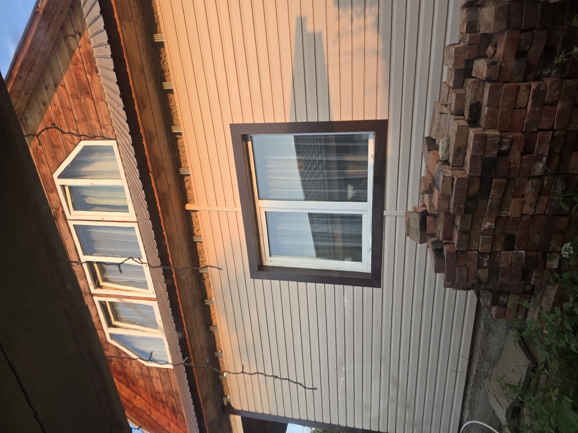 The image shows the exterior of a residential building under construction or renovation in Maksimiha, Russia. The building features a wooden upper section with a metal roof and several uniquely shaped windows, some of which are triangular. The lower section is clad in light-colored horizontal siding. A large, dark-framed window is visible on this lower level. In the foreground, a substantial pile of red bricks is stacked on the ground. Vegetation is present around the base of the building and the bricks. The lighting suggests it is either early morning or late afternoon, with the sun casting warm light and shadows. There are no people or animals visible in the image.