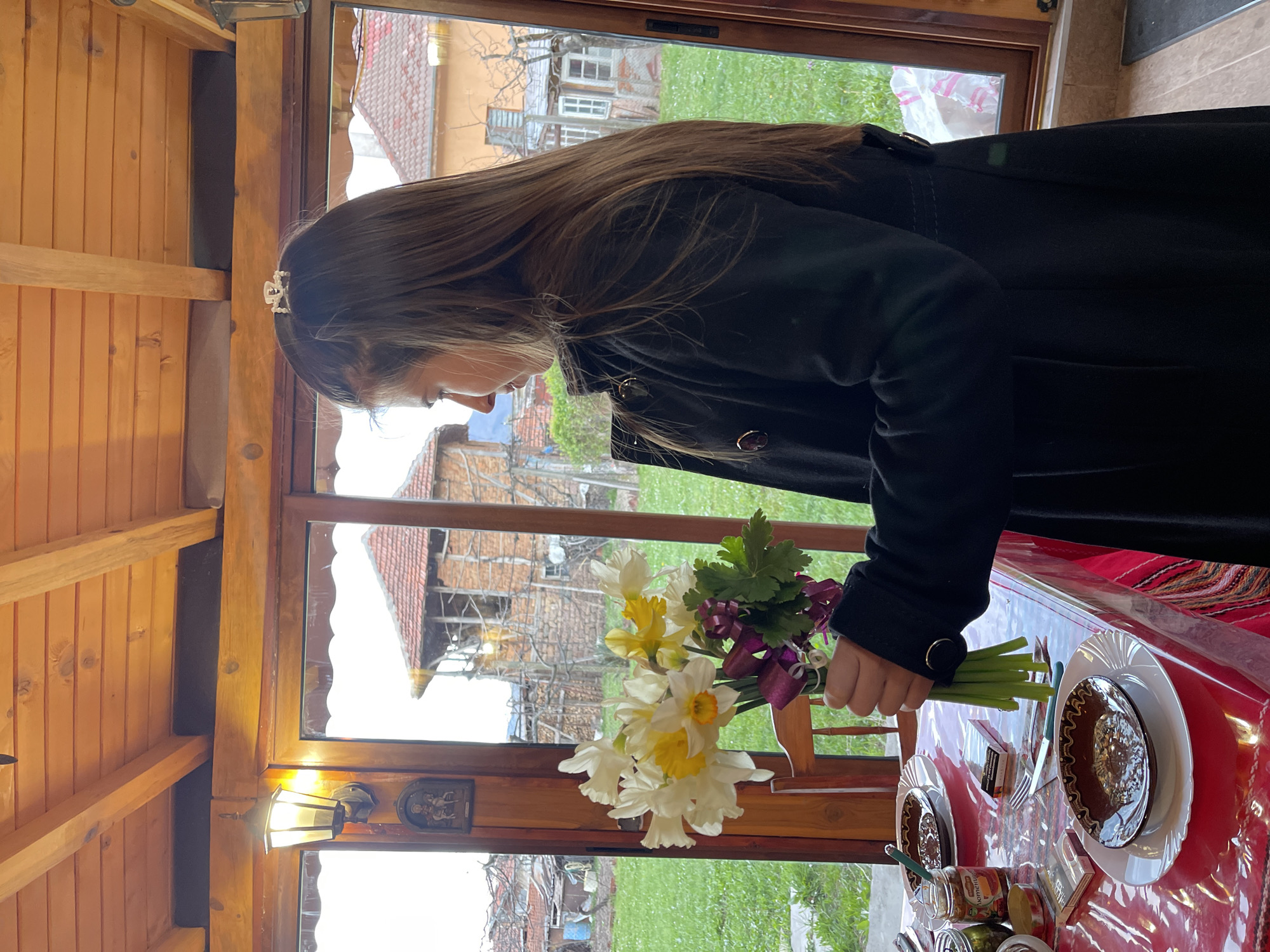 A person with long brown hair, wearing a black coat and a small decorative crown or headband, is holding a bouquet of daffodils and purple flowers in a wooden-framed structure, likely a veranda or enclosed patio, in Varshets, Bulgaria. Outside the window, there is a grassy yard with houses and structures of Varshets visible. Inside the room, a table is covered with a red and white patterned tablecloth set with plates and cutlery. Jars are visible on the table.
