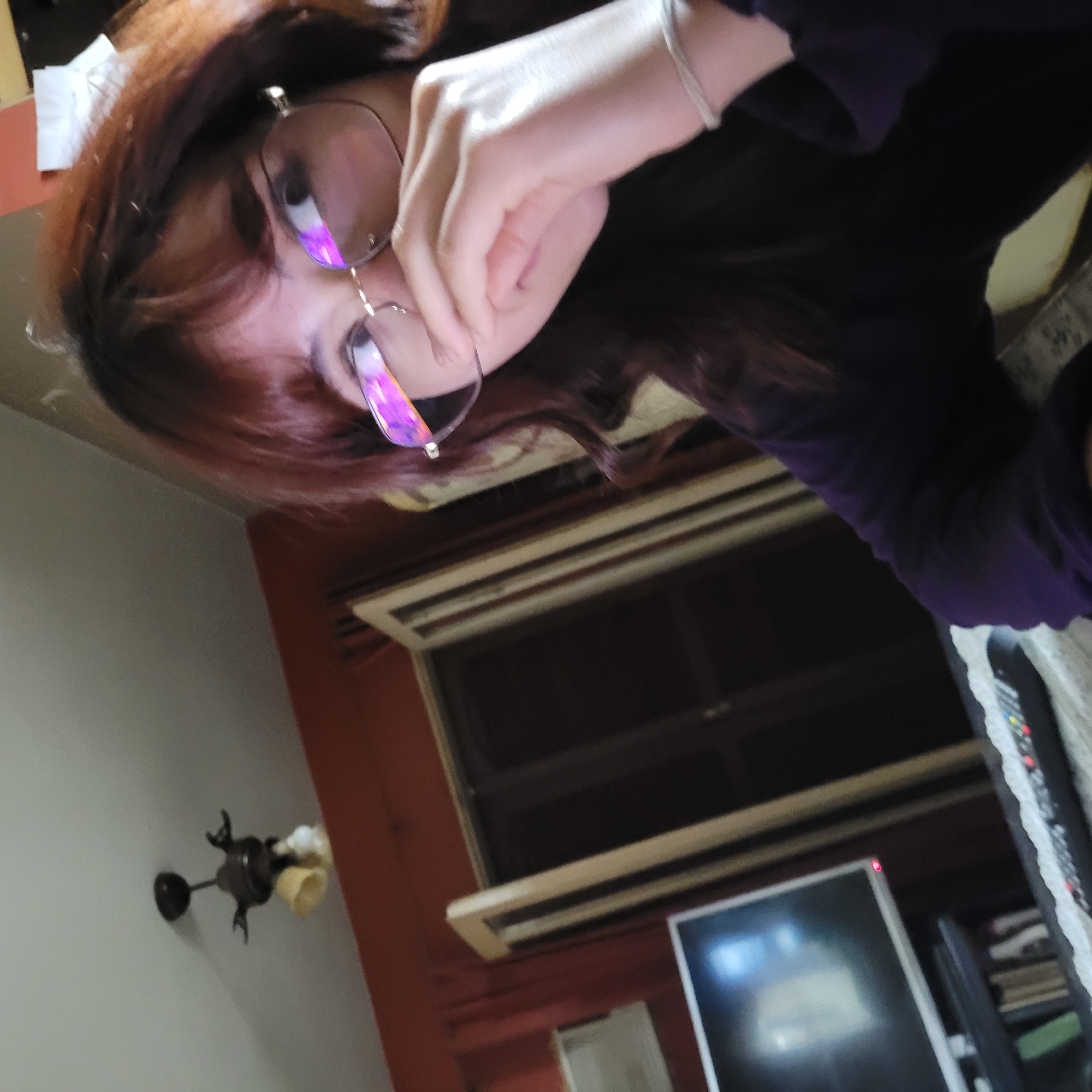 A tilted selfie shows a person with reddish-brown hair wearing glasses and a dark purple shirt. They are holding the glasses with their right hand. In the background, there's a window with closed blinds and a red wall. A dark-colored ceiling fixture is visible on a white wall to the left. A monitor and a remote control are also present in the lower portion of the image. The location appears to be indoors. I cannot confirm the location is the city of San Fernando del Valle de Catamarca, Argentina, because there are no defining features to do so.