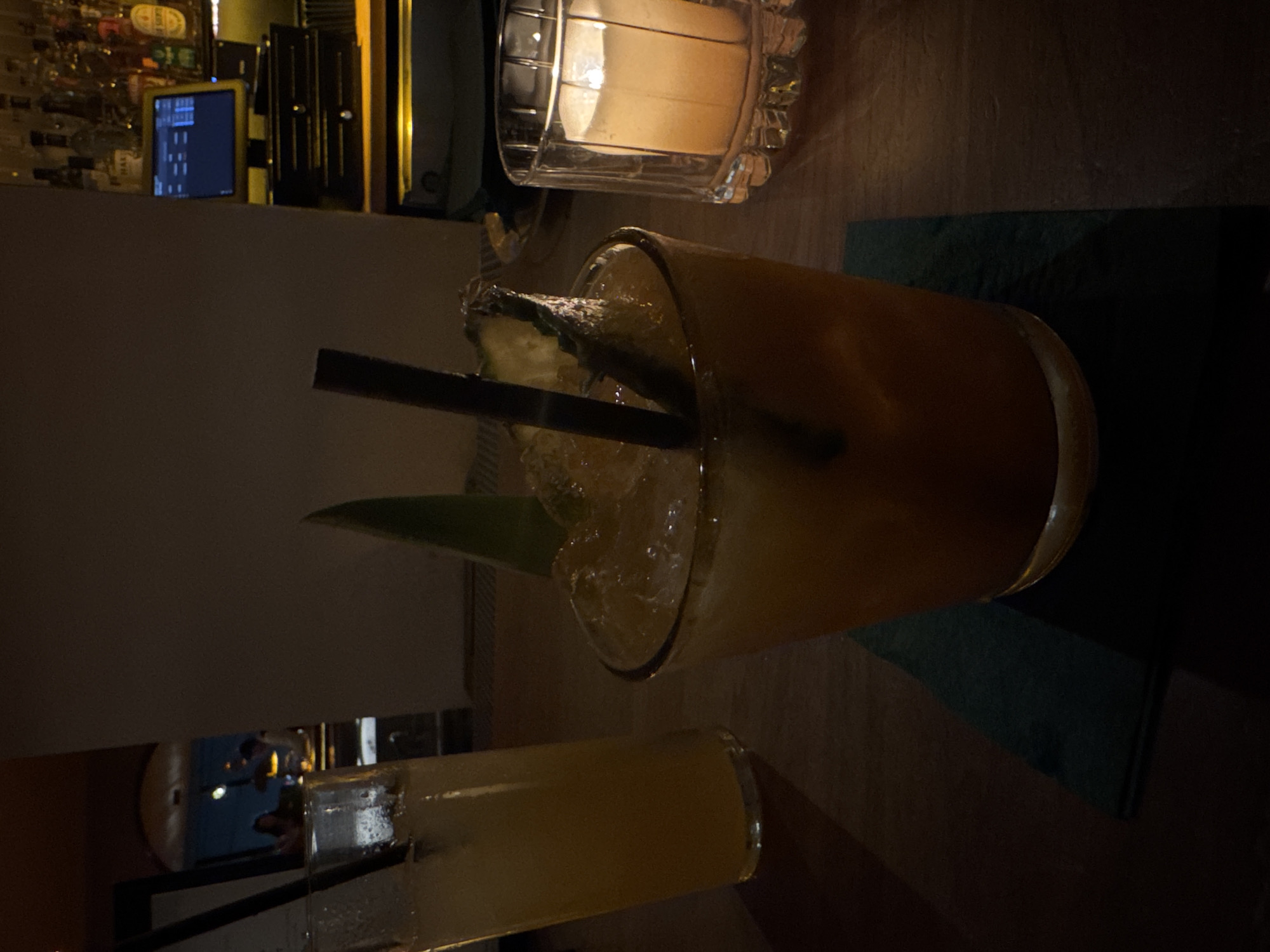 The image depicts a dimly lit, intimate setting, likely a bar or lounge in West Hollywood, United States. In the foreground, two cocktails are prominently displayed on a dark wooden surface. The main drink, positioned centrally, is an amber-colored beverage served in a short, wide glass filled with ice. It is garnished with a fresh pineapple wedge, a vibrant green leaf, and features a dark straw. Beneath this glass lies a dark green or blue napkin.
To the bottom left, a second, taller glass holds another amber-colored drink, also with ice and a dark straw, though only partially visible. A lit candle in an ornate, clear glass holder sits further back on the right, casting a warm, soft glow that illuminates the immediate area and contributes to the cozy atmosphere.
The background reveals elements typical of a bar. On the left, shelves stocked with various liquor bottles are visible, their labels indistinct due to the lighting and distance. Above the drinks, an electronic monitor or screen displays a dark interface with faint, unrecognizable blue or white text or graphics, possibly a point-of-sale system or digital menu. The overall ambiance suggests an evening or night out, with the focus on enjoying beverages in a relaxed environment. No people are clearly visible, though reflections in the background might hint at their presence. No legible text is visible.