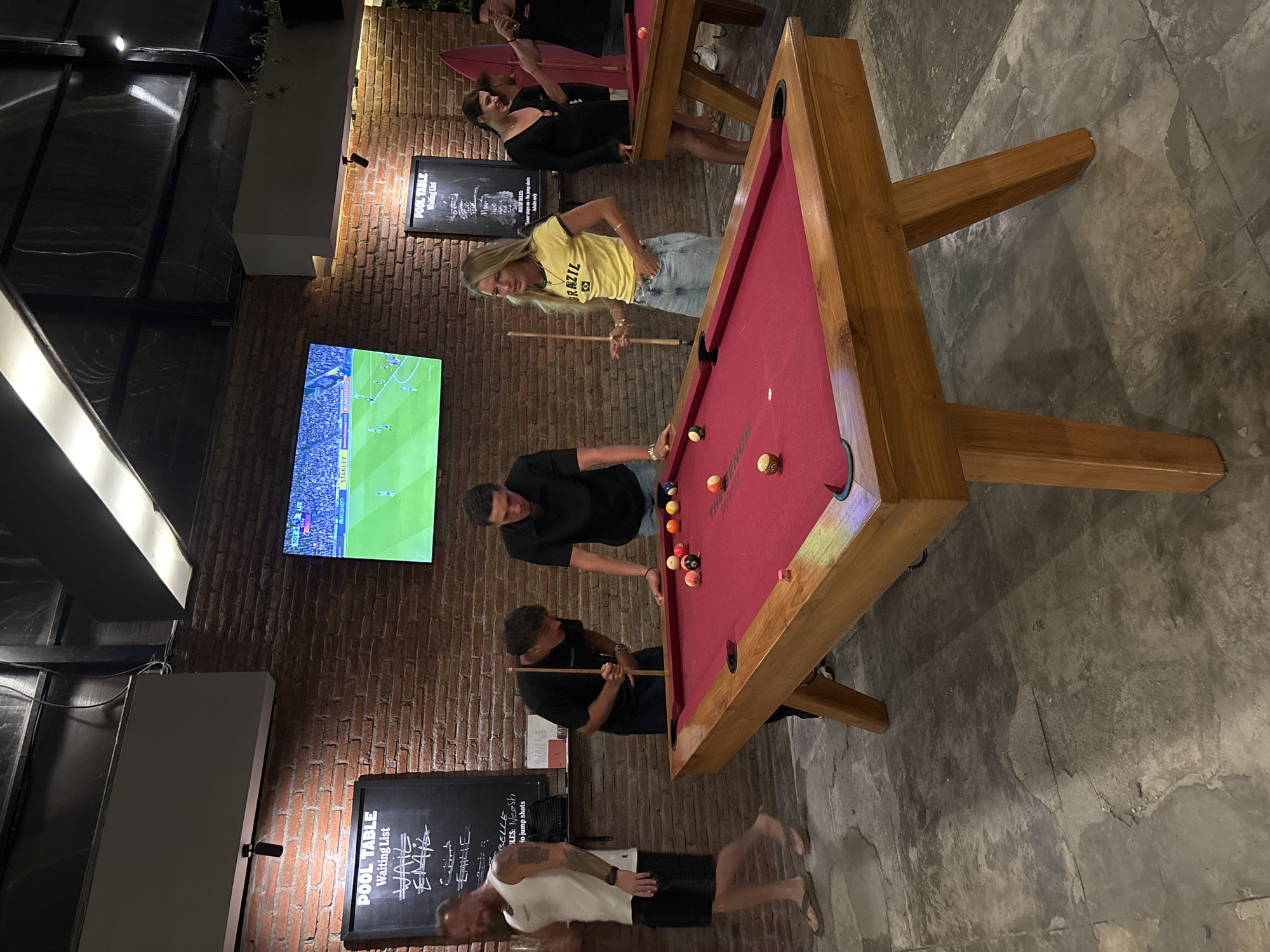 The image shows a casual scene at a bar in Kuta Selatan, Indonesia. Several people are playing pool at two tables. One pool table is triangular and red. There are at least four people playing at it. One woman in a yellow shirt is holding a pool cue. The second pool table is rectangular. On the wall are a TV showing a soccer match and a chalkboard labeled "POOL TABLE Waiting List". The bar has a brick wall and an exposed ceiling with artificial lighting. The floor is concrete.