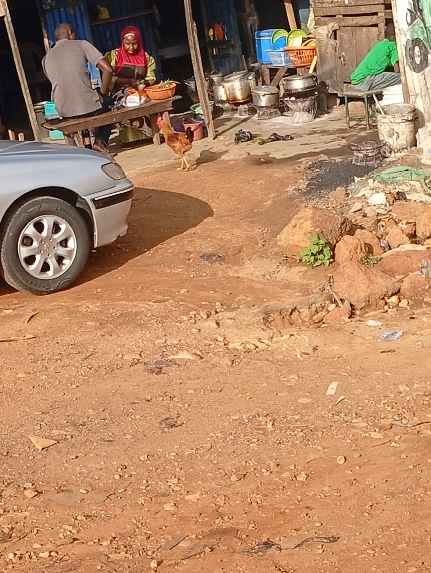 A chicken stands on a dirt path in front of a modest outdoor food stall in Rafin Par, Nigeria. The stall features a wooden bench where a man in a grey t-shirt and jeans sits, facing away from the camera. Next to him, a woman wearing a red headscarf and a green and yellow patterned garment is seated and appears to be preparing food on the same bench. In the background, various cooking pots are visible on small stoves, along with stacks of plastic containers and baskets. A portion of a silver car is in the foreground on the left, with its tire prominently displayed. The ground is a mix of dirt and scattered stones, suggesting a somewhat unpaved urban or semi-urban environment.