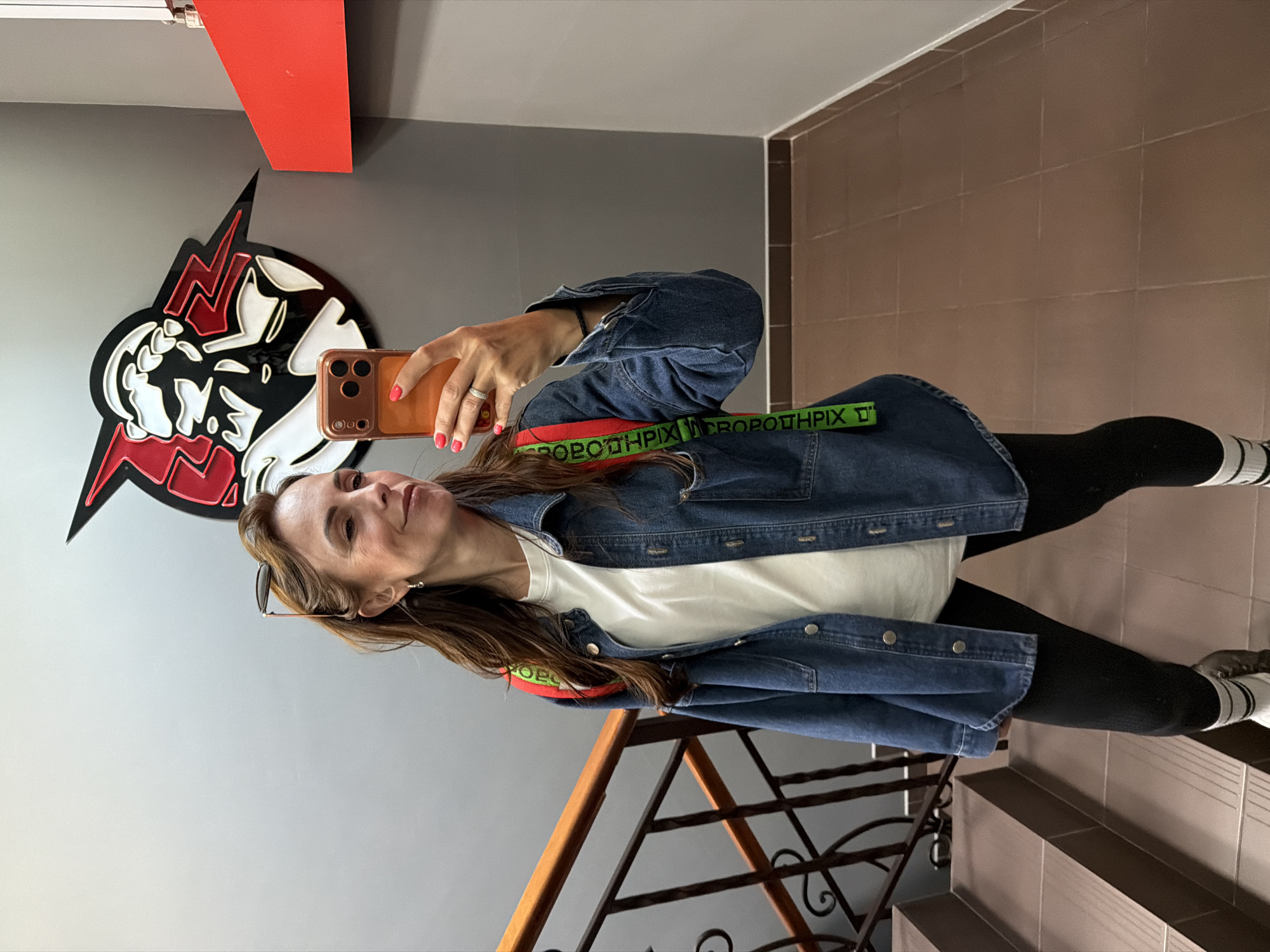 A woman takes a mirror selfie in a hallway with a staircase on the left and a gray wall with a red and black logo on the right. She is wearing a denim jacket over a white t-shirt, black leggings, and socks with stripes. A phone is held in her right hand, and a backpack with red straps and a green strap with text is visible on her shoulders. The logo on the wall appears to be a stylized character with lightning bolts. The lighting is artificial and bright. The floor is tiled in a reddish-brown color. The environment appears to be indoors, possibly a public building or a gym. The location is not specified in the image itself, and there is no visual information to confirm it is in Birobidzhan, Russia.