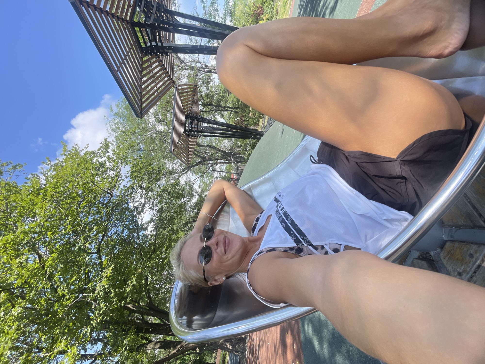 In Novorossiysk, Russia, a blonde woman with sunglasses and a white top with leopard print accents is lying on a slide in a park. She is holding her head with one hand and smiling at the camera. The slide is made of metal and green plastic. In the background, there are trees and a blue sky with clouds. There are also some modern structures with wooden slats overhead. The ground is covered with a red and green rubberized surface.