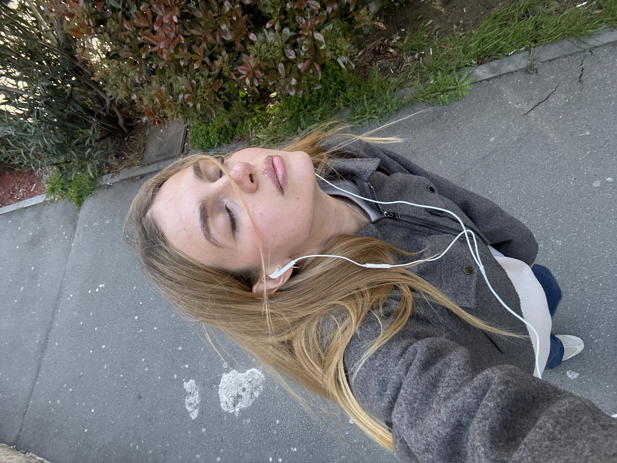 A woman lies on a gray asphalt sidewalk in Tbilisi, Georgia. She is fair-skinned, with long blonde hair and closed eyes, wearing a gray wool coat over a white shirt and blue pants. White earphones are in her ears and the cord runs across her coat. Patches of green grass and shrubs are visible along the edge of the sidewalk. A bush with reddish-brown leaves is visible behind her. The angle of the photograph suggests it was taken from an elevated perspective. The overall scene suggests an unconscious or unwell person in a public space.