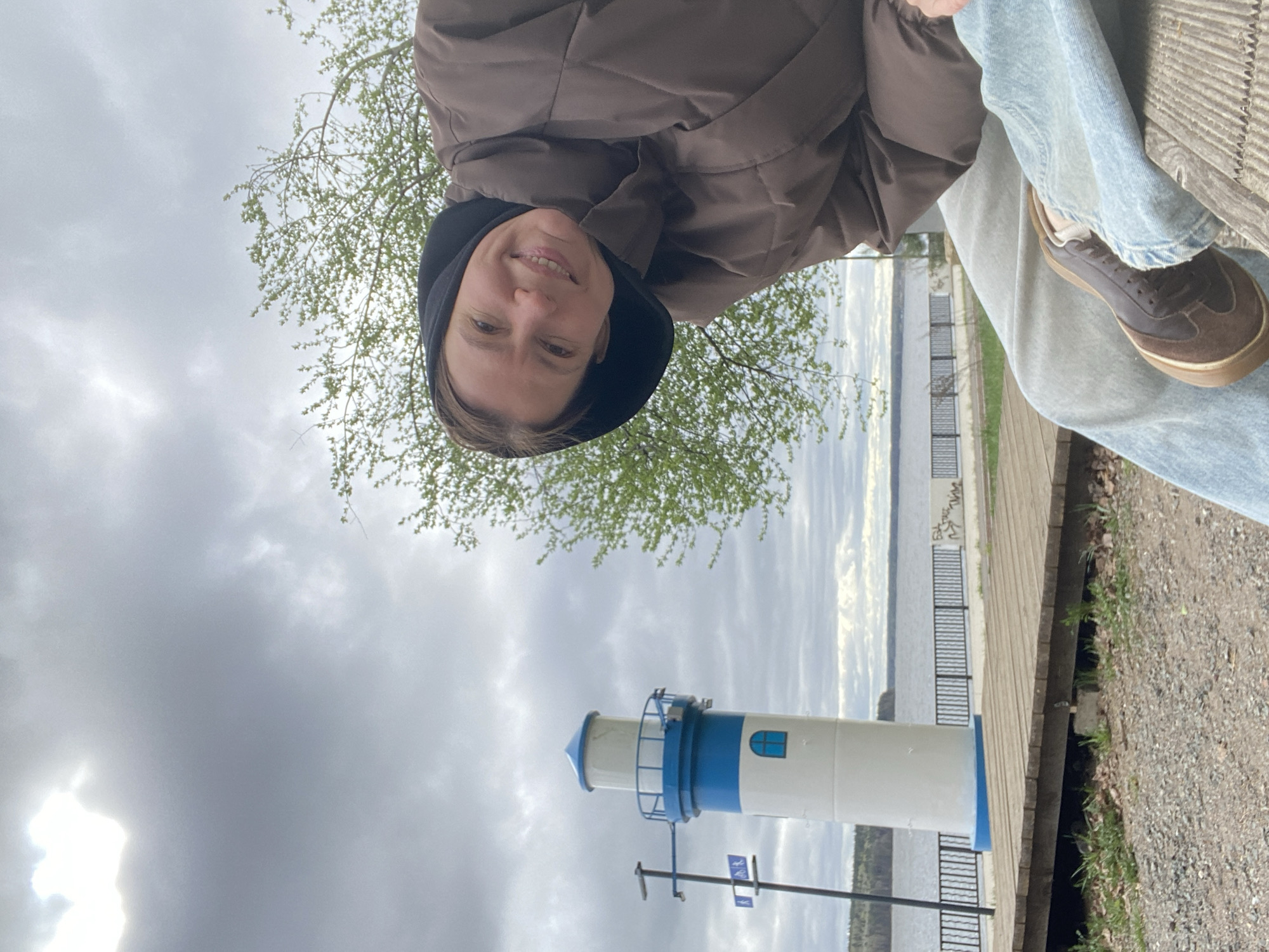 A selfie-style image features a woman with fair skin and short brown hair, wearing a brown hooded jacket and light blue jeans. She is smiling at the camera. The photo is taken at an odd angle, seemingly looking upwards.
In the background, a white and blue lighthouse is visible against a cloudy sky. Also visible are trees, a body of water, and what appears to be a walkway or path.
The location is Chaikovsky, Russia.