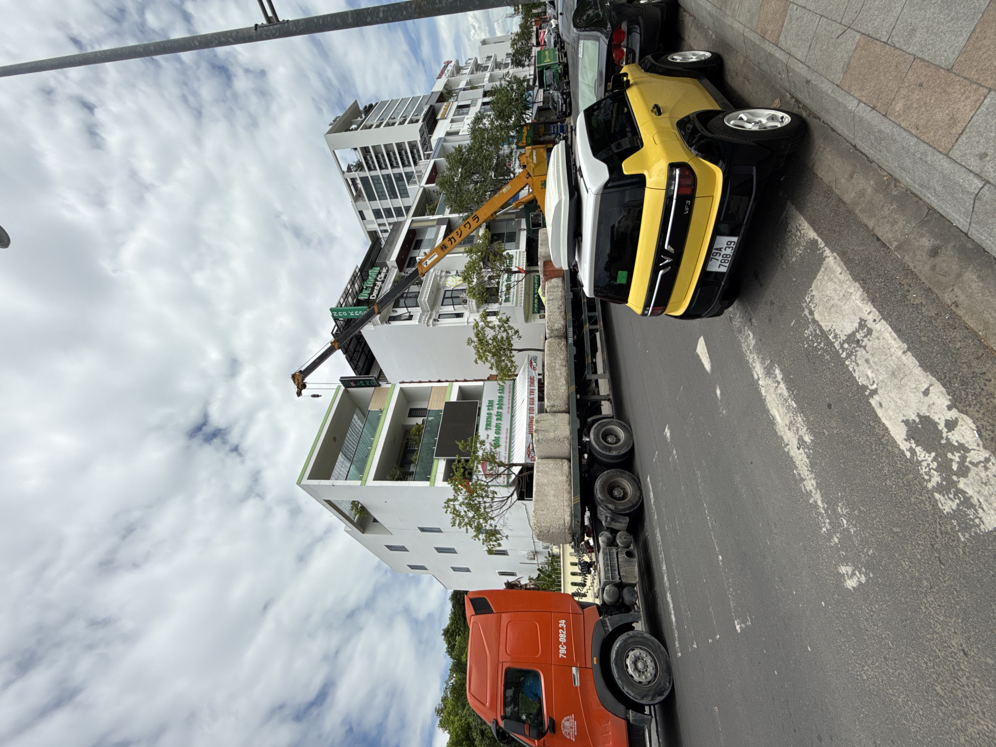 An orange semi-truck is parked on a street in Nha Trang, Vietnam, with its trailer loaded with large, cylindrical concrete blocks. A yellow electric car with a white roof box is parked behind the truck. In the background, a crane is positioned over the trailer, and several multi-story buildings line the street. The sky is cloudy.