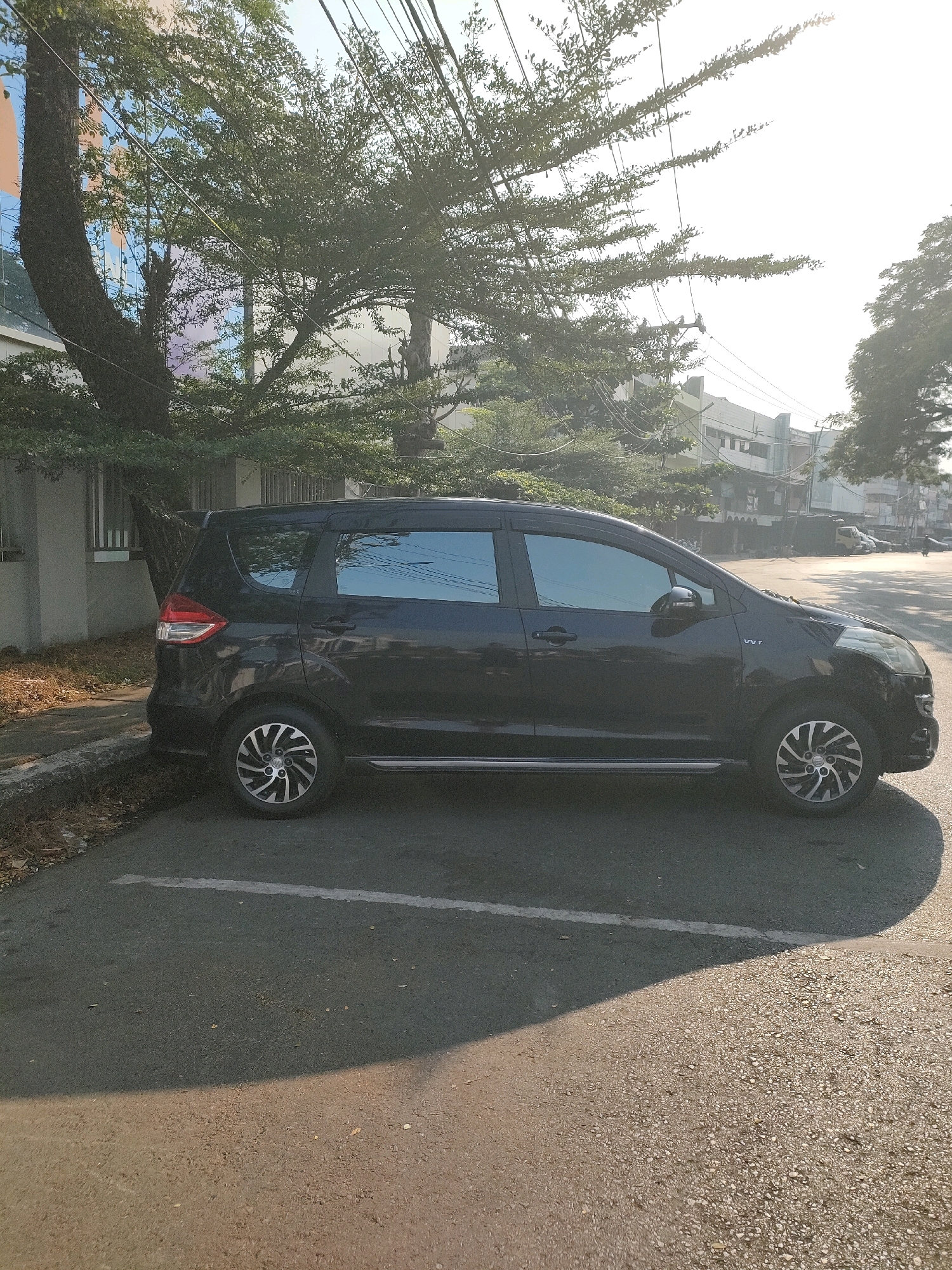 A black Suzuki Ertiga is parked on the side of a street in Banjarmasin, Indonesia. The car is facing the right side of the image, and its windows are tinted. The vehicle is parked along a curb, with fallen leaves and debris on the ground next to it. In the background, there is a sidewalk, a building with windows reflecting the sky, and lush green trees with thin branches. Electrical wires crisscross above the trees. The street appears to be paved, with a white line marking a parking space. The sun is shining brightly, casting a long shadow of the car onto the road. In the distance, other buildings and vehicles are visible, suggesting an urban environment. The sky is bright and hazy, typical of a sunny day in a tropical climate.