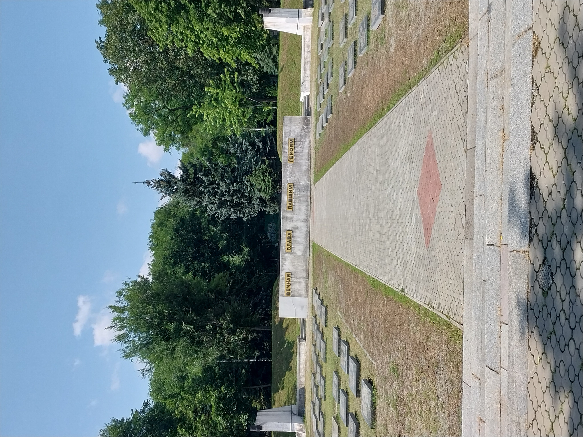 The image depicts a memorial park with rows of grey rectangular grave markers on a grassy area. A paved pathway leads towards a concrete wall with yellow text. The wall has inscriptions in Cyrillic that translate to "Eternal Glory to Our Heroes." The background consists of lush green trees under a clear blue sky with a few scattered clouds. Two white obelisk-like monuments flank the wall, one on each side, and a dark spire stands to the left of the left obelisk. This memorial is located at the Dnipro memorial complex of the Fallen Defenders of the Fatherland in Dnipro, Ukraine. There are no people visible in the image.