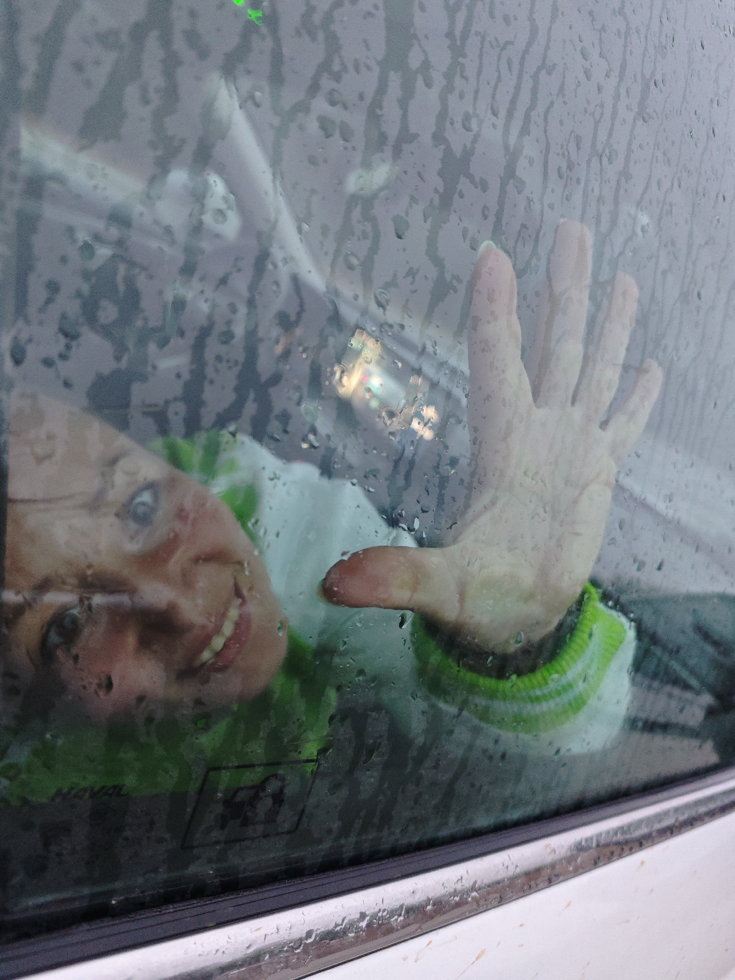 A woman with visible teeth and eyes is partially seen through a car window covered in raindrops. Her hand is pressed against the glass. A green and white striped garment is visible around her neck. The window also reflects a blurry light source. The car's exterior is white. A window sticker of a horse and a horse rider is near the bottom of the frame. The location is Dubovaya, Russia.