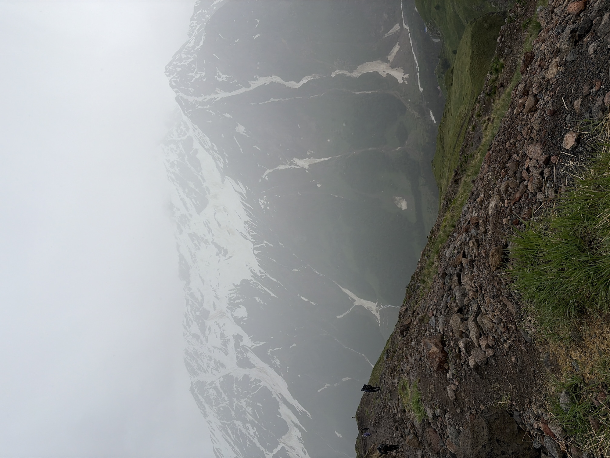 The image depicts a group of hikers on a rugged, rocky mountain trail. In the background, a vast, snow-capped mountain range is shrouded in mist. The foreground shows a steep incline covered in loose stones and patches of grass. Several individuals, identifiable by their backpacks and trekking attire, are walking along the path. One hiker is positioned closer to the camera, facing away from it, while others are further up the trail, spread out as they ascend. The environment suggests a challenging and remote mountain terrain, consistent with the high-altitude landscapes around Terskol, Russia, which is known for its proximity to Mount Elbrus. The overall atmosphere is overcast and slightly hazy, obscuring distant details and contributing to a sense of vastness and elevation.