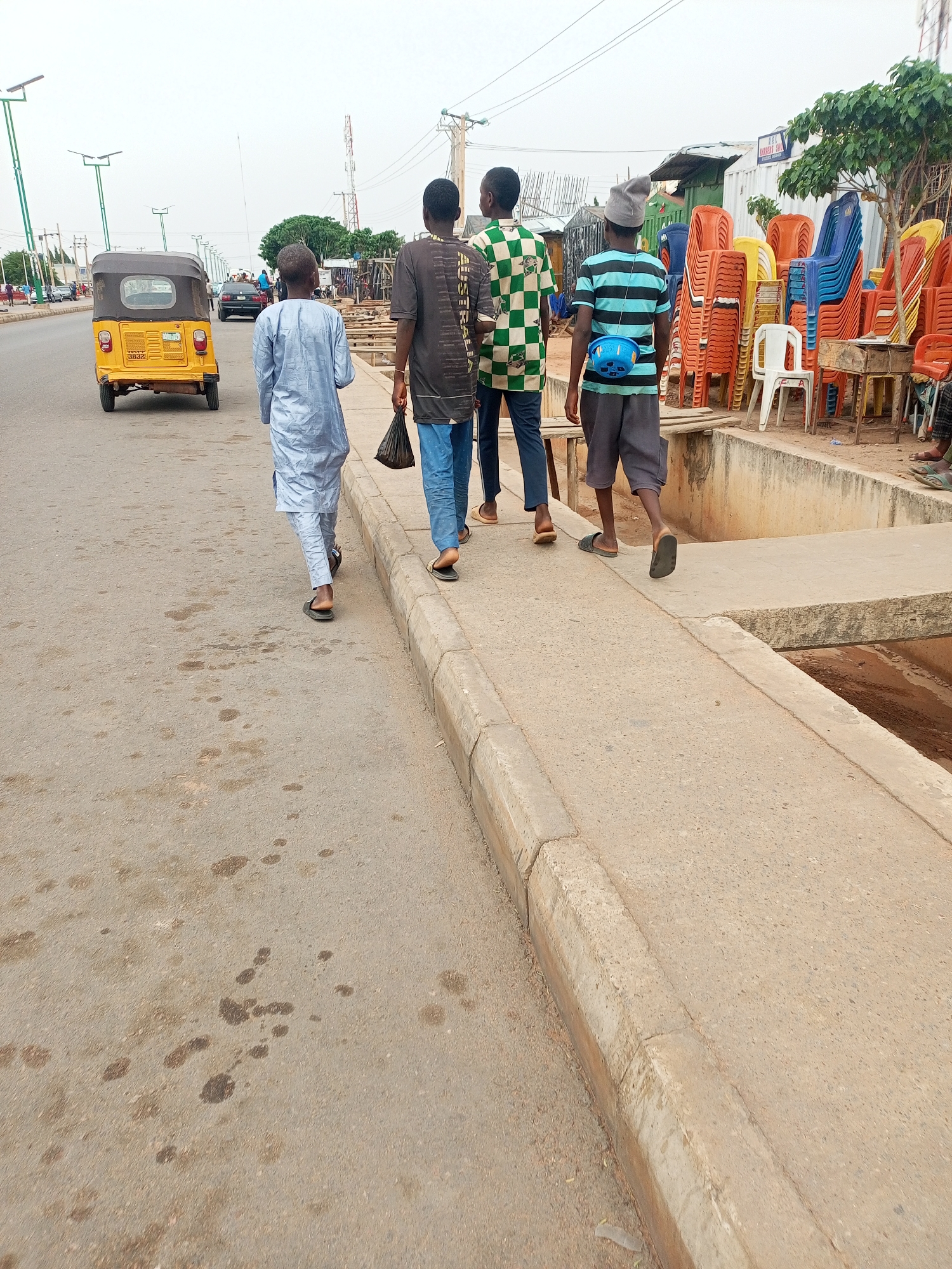 Four people walk together down a concrete sidewalk, approaching a pedestrian bridge over a drainage ditch. The person farthest to the left wears a light blue robe, while the other three wear casual clothes. The person farthest to the right wears a gray hat and has a small blue bag slung across their chest. To the right, stacks of orange, yellow, blue, and white plastic chairs sit on a table and the ground next to a shop. Across the road to the left, a yellow tricycle taxi drives away from the camera. The asphalt of the road has dark stains. The sky is overcast. Based on the environment and architecture, it's likely this image was taken in a city like Tempure, Nigeria.