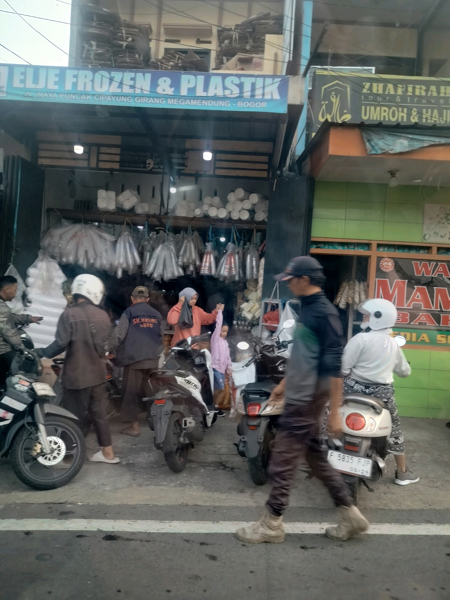 The image depicts a street scene in front of storefronts in Megamendung, Indonesia. A prominent blue sign reads "ELJE FROZEN & PLASTIK" with smaller text below indicating "JL. RAYA PUNCAK CIPAYUNG GIRANG MEGAMENDUNG - BOGOR." To the right, a yellow sign with Arabic calligraphy and text in Latin characters says "ZHAFFIRAH TOUR & TRAVEL" and "UMROH & HAJI."
Several people are visible. A person wearing a dark jacket and a white helmet stands near a parked motorcycle. Another person in a brown jacket and white helmet is seen from the back. Further to the right, a person in an orange hijab and a pink shirt holds hands with a young person in a pink shirt and blue shorts. A person in a black long-sleeved shirt and a black cap is walking across the foreground, with a blurred white helmet on their head. Another individual in a white long-sleeved shirt and white helmet is seated on a scooter.
Various objects are present. Multiple motorcycles are parked along the street. Storefronts display products, including stacked white containers, rolled white materials, and hanging plastic items. A green tiled wall is visible on the right side of the image. The ground is paved asphalt with white painted lines. The overall atmosphere suggests a busy commercial area.