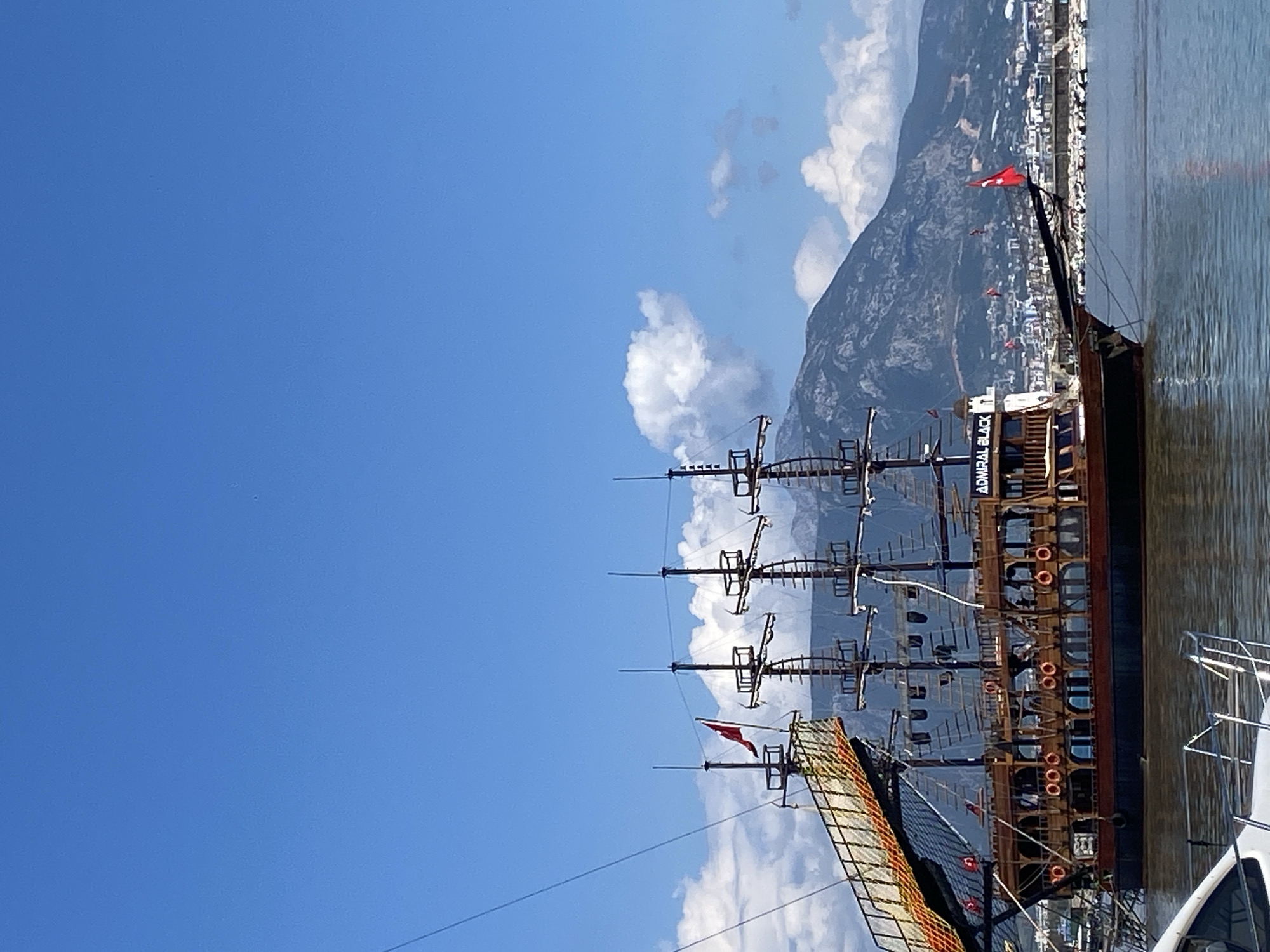 The image captures a docked pirate ship named "Admiral Black" in Alanya, Turkey. The ship has multiple masts and is flying red flags. Behind the ship, a mountain range is visible, and a city skyline is situated along the waterfront. The sky is blue with scattered clouds. To the right of the pirate ship, a smaller, white boat is also docked. The scene appears to be taken during a sunny day.
