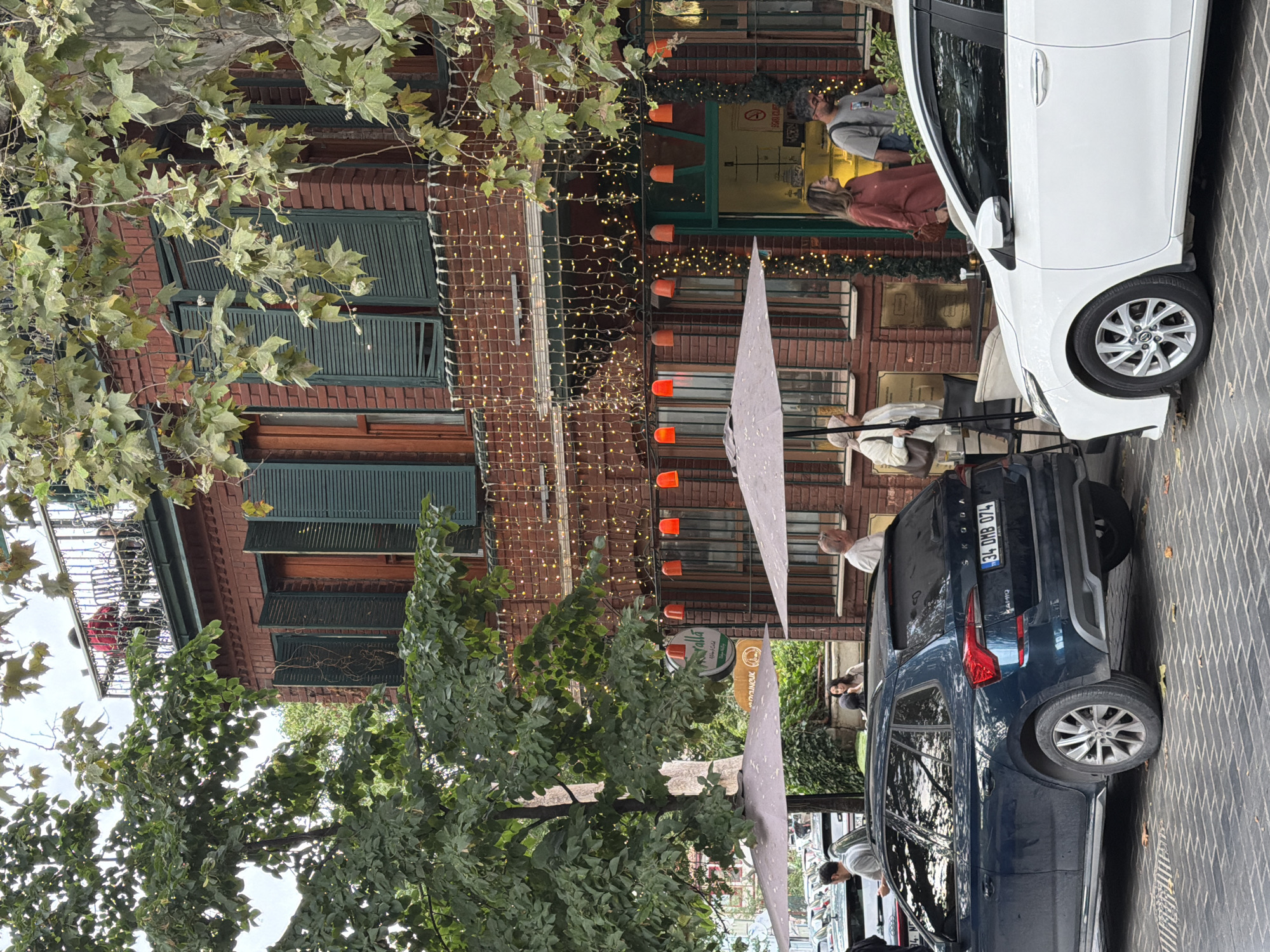 The image captures a street scene in Üsküdar, Turkey, in front of a multi-story brick building adorned with green shutters and string lights. A tree with green leaves partially obscures the upper floors. Two cars are parked on a cobblestone street; a dark blue Skoda is in the foreground on the left, and a white sedan is on the right. Several people are visible. On the balcony of the building, two individuals are seated. On the ground level, patrons are seated at outdoor tables under umbrellas, and others are walking on the sidewalk in front of the building's entrance. Signs are mounted on the building's facade, one appearing to be a menu or establishment name with green accents. The overall atmosphere suggests a commercial area with outdoor dining or cafe seating.
