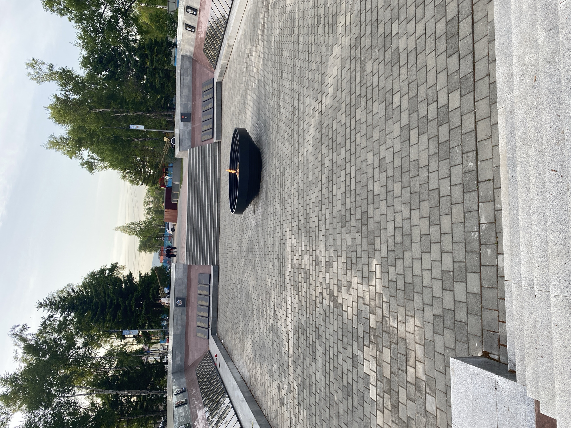 The image depicts a view of the "Eternal Flame" monument in Artyom, Russia. The flame burns within a large, dark, circular bowl set on a paved plaza. The plaza consists of gray, rectangular paving stones laid in a regular pattern. Steps lead down from the plaza to the left, flanked by stone walls adorned with plaques and images, likely commemorating historical events or figures. Beyond the wall and stairs are trees and a glimpse of the city. To the right of the plaza, a set of stairs descends towards the foreground. The sky is overcast.