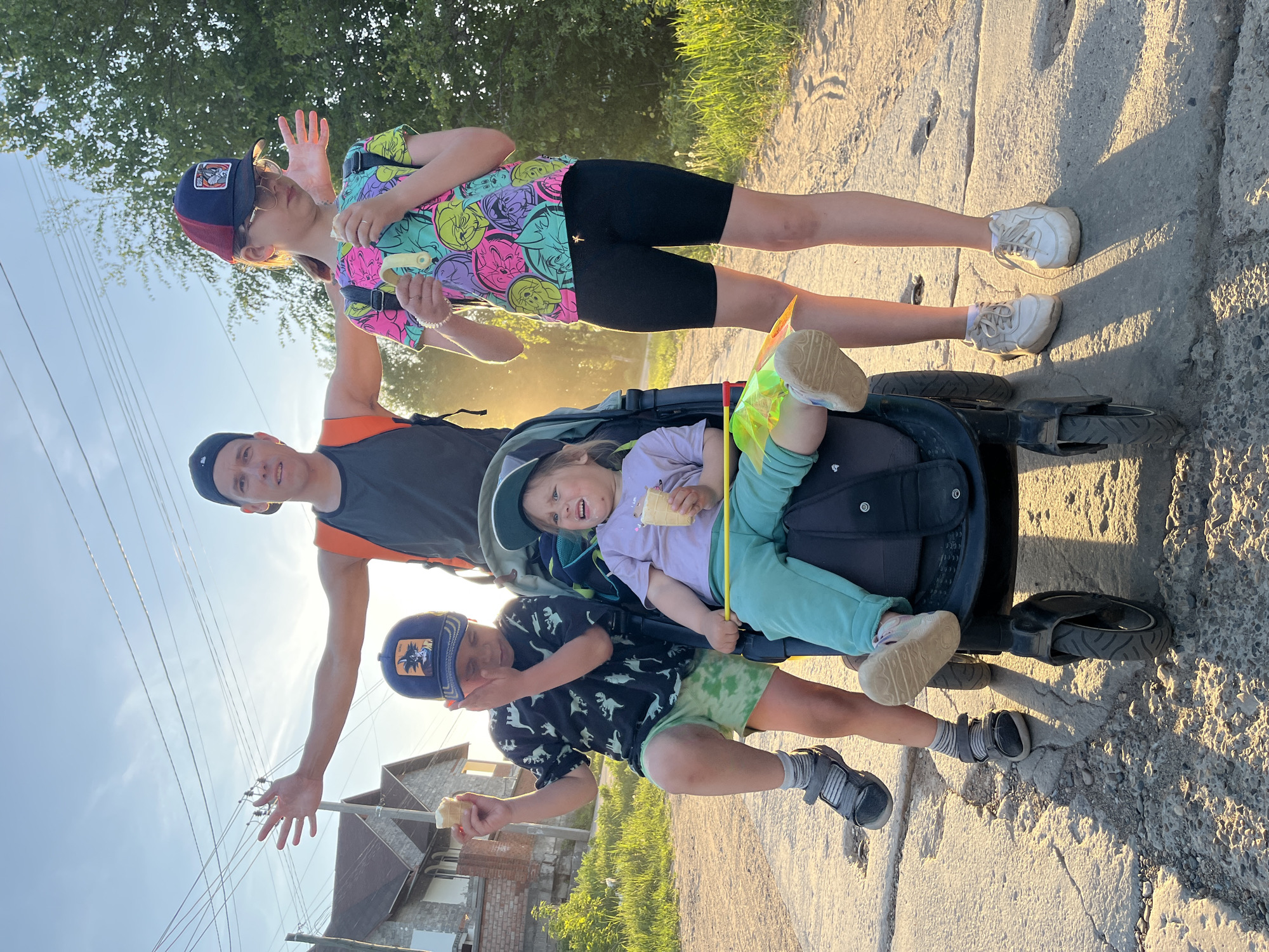 A group of four people is posing on a paved road or pathway in Chaikovsky, Russia. From bottom to top, there is a boy in a dinosaur print shirt and green shorts wearing a baseball cap and grey athletic shoes holding an ice cream cone. Next is an adult with short brown hair wearing an athletic shirt with orange shoulder stripes and black cap. A young child in a purple shirt and light blue pants sits in a stroller. They are holding a small ice cream cone and a pinwheel. At the top of the photo, another child in a t-shirt featuring Disney cartoon characters with black biker shorts, white sneakers and a baseball cap is holding an ice cream cone. All four individuals are smiling or laughing while posing for the photo. The background includes a house, trees, and a partially visible blue sky with setting sun.