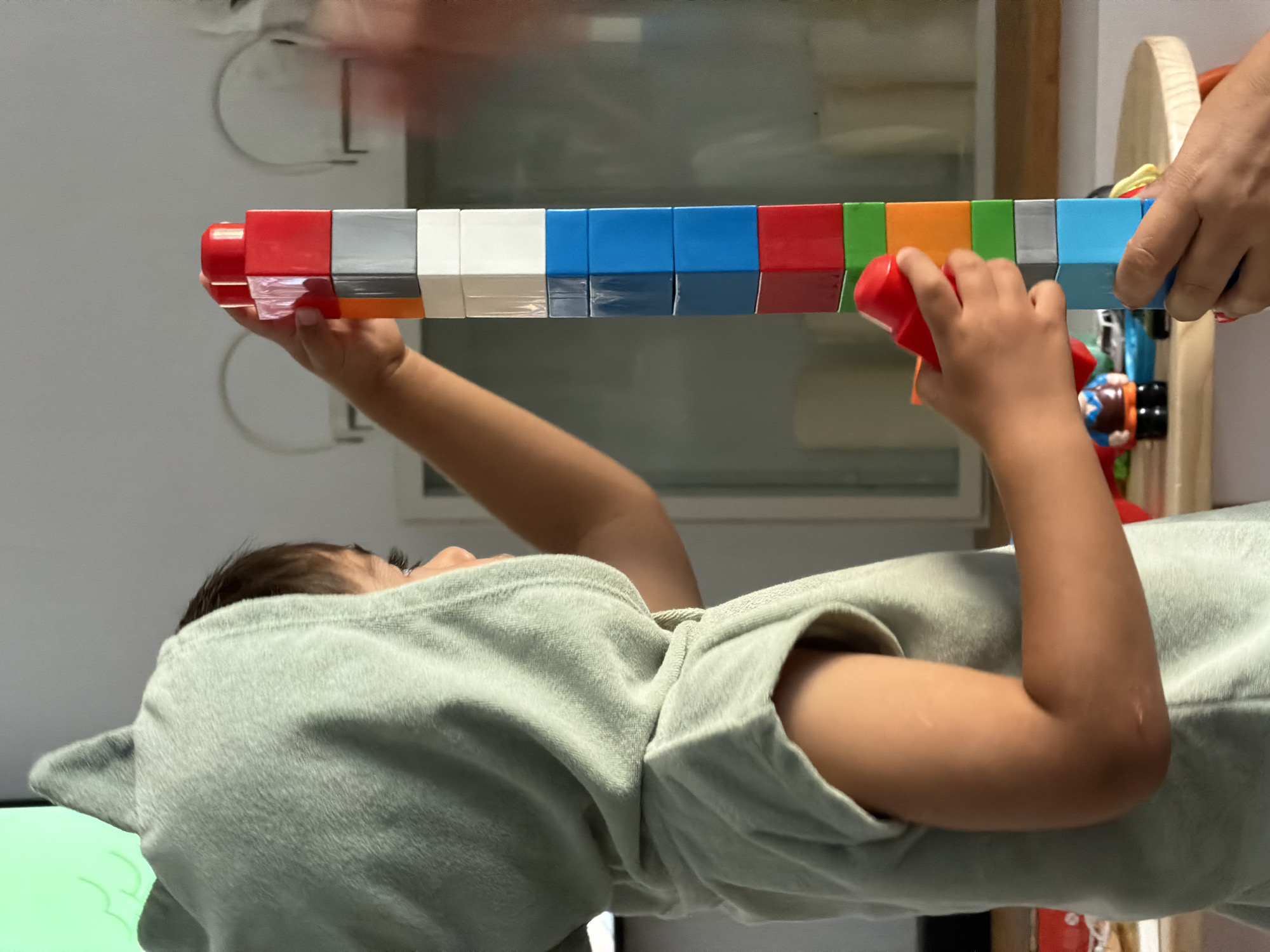 In a domestic indoor setting, likely a living room or playroom, a young child wearing a light green hooded garment with shark fin detailing on the hood is actively engaged in constructing a tall tower of colorful interlocking blocks. The child's right hand is positioned to place a red block at the top of the tower, while their left hand supports the structure. The tower itself is composed of blocks in various colors including red, white, grey, orange, blue, and green. In the background, a blurred suggestion of furniture and a window is visible. The overall scene captures a moment of focused play and creativity. The image does not provide any specific details about Junglinster, Luxembourg, as the location is not visually represented or indicated within the provided visual information.