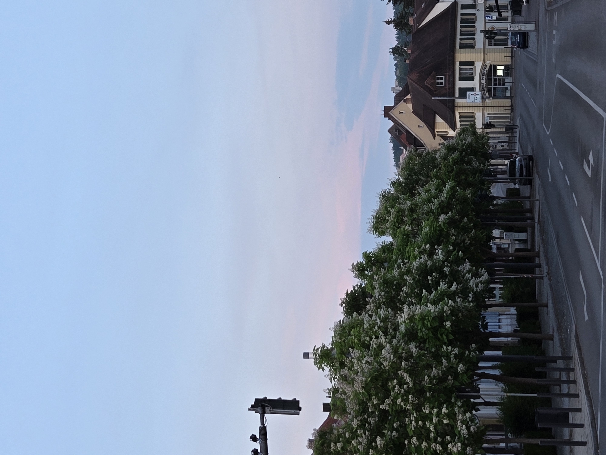 The image depicts a street scene at dusk in Ansbach, Germany, specifically on Johann-Sebastian-Bach-Platz. A traffic light stands prominently on the left side of the frame, over a row of trees with white blossoms. The street is lined with buildings, one of which has the sign "GERBERT PFEUFFER" above its entrance. Two cars are parked on the right side of the street, and one is parked on the left. The sky is a soft gradient of blue and pink, indicating the end of the day. The overall impression is one of a quiet, orderly urban environment.