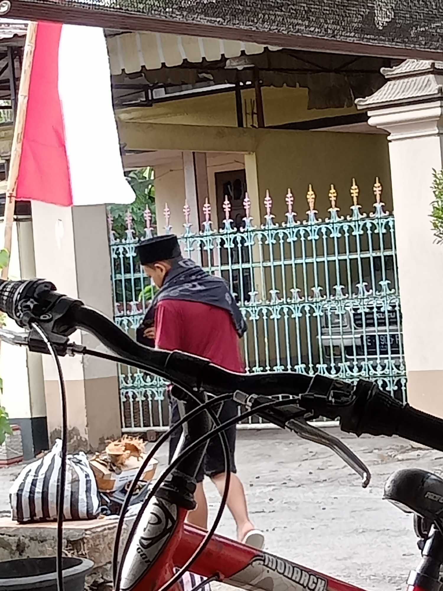 In Simo, Indonesia, an individual wearing a red shirt, black peci hat, and a dark patterned scarf walks away from the camera. They are positioned in front of a light green building with a decorative green fence. To the left, a red and white Indonesian flag hangs from a pole. In the foreground, a bicycle's handlebars and part of the frame are visible. Next to the bicycle, a striped bag and some boxes are on the ground.