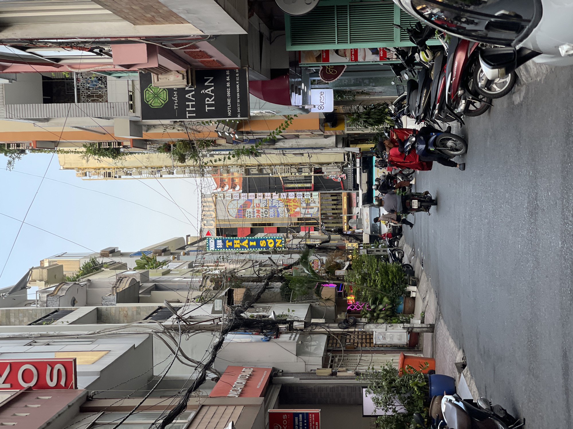 The image captures a street scene in the city of Thủ Đức, Vietnam. The narrow street is lined with buildings of varying heights and architectural styles. Power lines crisscross overhead. Several motorbikes are parked along the edge of the street. Pedestrians and vehicles move along the street. The lighting suggests it is daytime. The atmosphere appears busy and urban.