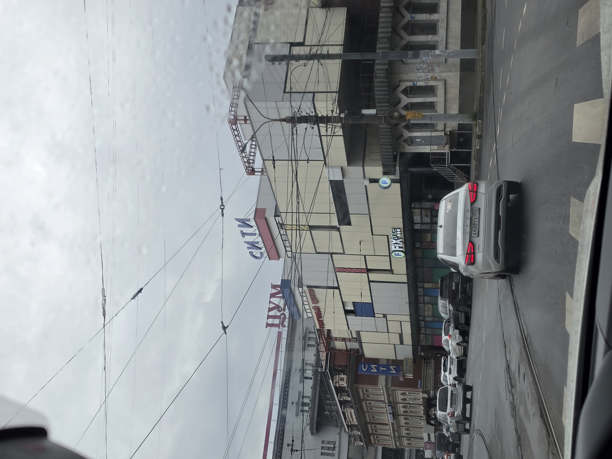A grey, overcast day is depicted in a city street scene in Nizhny Novgorod, Russia. The image captures a view from a vehicle looking forward and slightly to the right. In the foreground, the wet asphalt street shows lane markings and a pedestrian crossing. Multiple cars are visible on the road; a white sedan with the license plate "A438TX 252" is prominently in the right lane, and other vehicles are in traffic ahead.
Dominating the background is a large, multi-story building with a modern, somewhat abstract facade, featuring a pattern of cream and beige panels of varying sizes, some with red accents. A sign with blue Cyrillic lettering reads "СИТИ" (SITI). Below this, another sign in red Cyrillic lettering spells "ЦУМ" (TSUM). To the left of this structure, a more traditional building with red brick and detailed white trim is visible, also bearing a blue "СИТИ" sign and "ТОРГОВЫЙ ЦЕНТР" (Shopping Center).
The scene is heavily crisscrossed with numerous overhead wires, likely for public transport like trolleys or trams, and electrical infrastructure. A street lamp is positioned in front of the modern building. The overall impression is of an urban commercial district on a damp day, with a mix of architectural styles and active traffic.