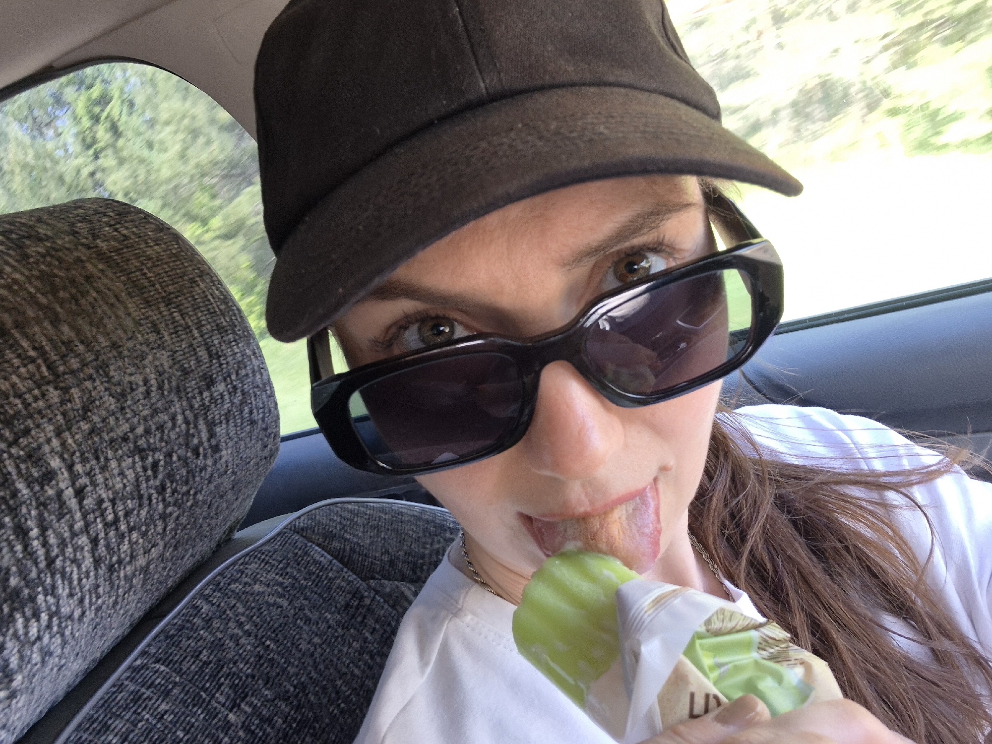 A selfie taken inside a vehicle shows a person wearing a black baseball cap and black sunglasses, sticking their tongue out to lick a green popsicle. The person is wearing a white t-shirt. A gray upholstered seat is visible to the left. A blurred view of trees is visible through the vehicle's window in the background. The location is Unknown, Unknown.