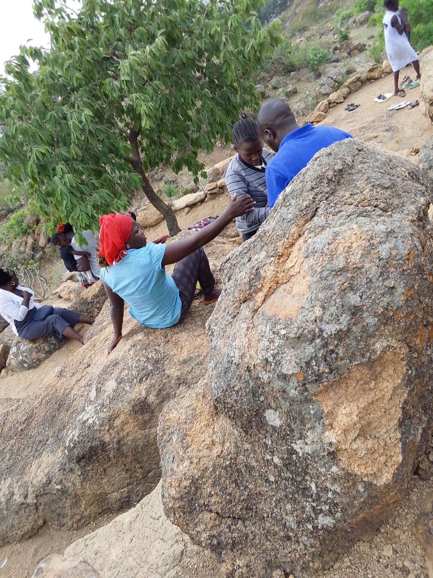 The image depicts a group of people gathered on a rocky, sandy hillside in what appears to be a natural setting. A large tree is located to the left of the frame. In the foreground, a woman wearing a red head covering and a blue shirt is seated on the rock, with her arm extended. Near her, another woman in a white shirt and dark trousers is sitting. Further up the slope, a couple is standing, with the man wearing a blue shirt. Another person is seen in the background wearing a white dress. Several pairs of sandals are scattered nearby, suggesting a relaxed atmosphere. The ground is covered in sand and small rocks, and the vegetation is sparse. The location is Gura Top, Nigeria.