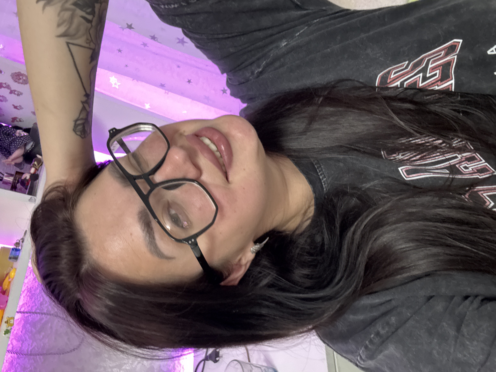 The image shows a woman indoors, with a shelf behind her containing various items, possibly in a bedroom or living area. The lighting is predominantly purple, suggesting decorative lights or a light fixture. The woman is wearing glasses and a dark t-shirt with a graphic design. She is looking towards the camera and appears to be posing. There is a tattoo visible on her arm. The background includes a patterned curtain. No specific details indicate the location being in Tolyatti, Russia.
