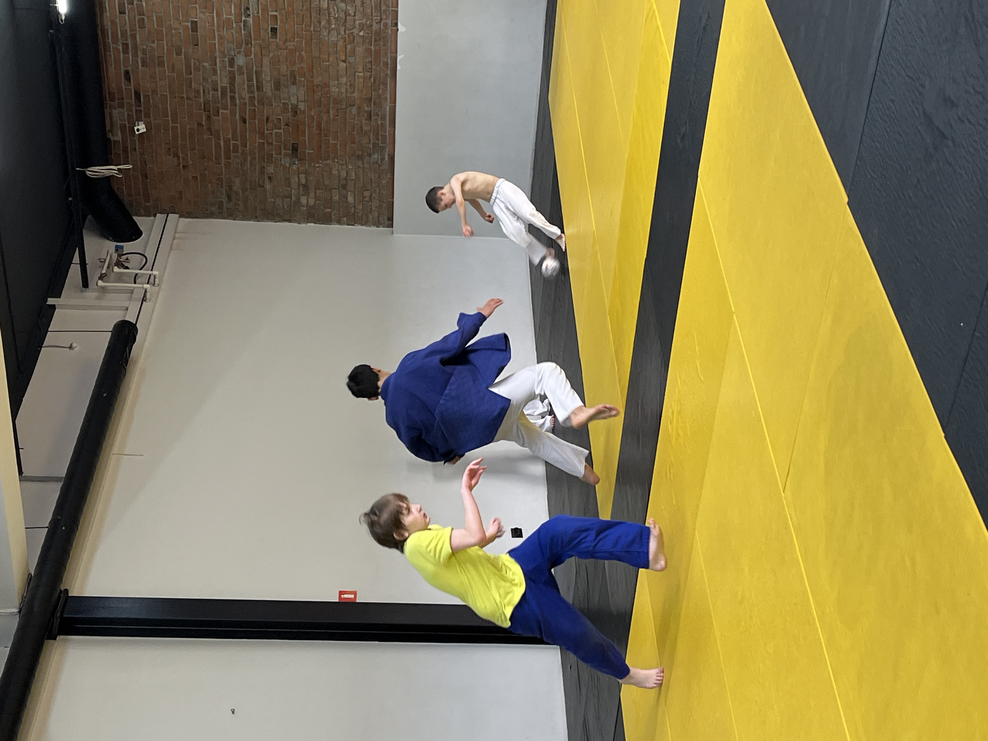 The image captures a scene from what appears to be a martial arts practice, possibly Judo, taking place in a gym or training area. Three individuals are visible, two wearing Judo uniforms (judogi). One person is dressed in white pants and is shirtless. A person in a yellow shirt and blue pants is near the bottom of the shot. The others wear Judo gi, blue and white. All of the people appear to be suspended in the air. The background includes a brick wall, a white wall, and what appear to be floor mats with yellow and black stripes. The location is identified as Krasnoyarsk, Russia. The scene appears to capture a moment of physical activity or training.