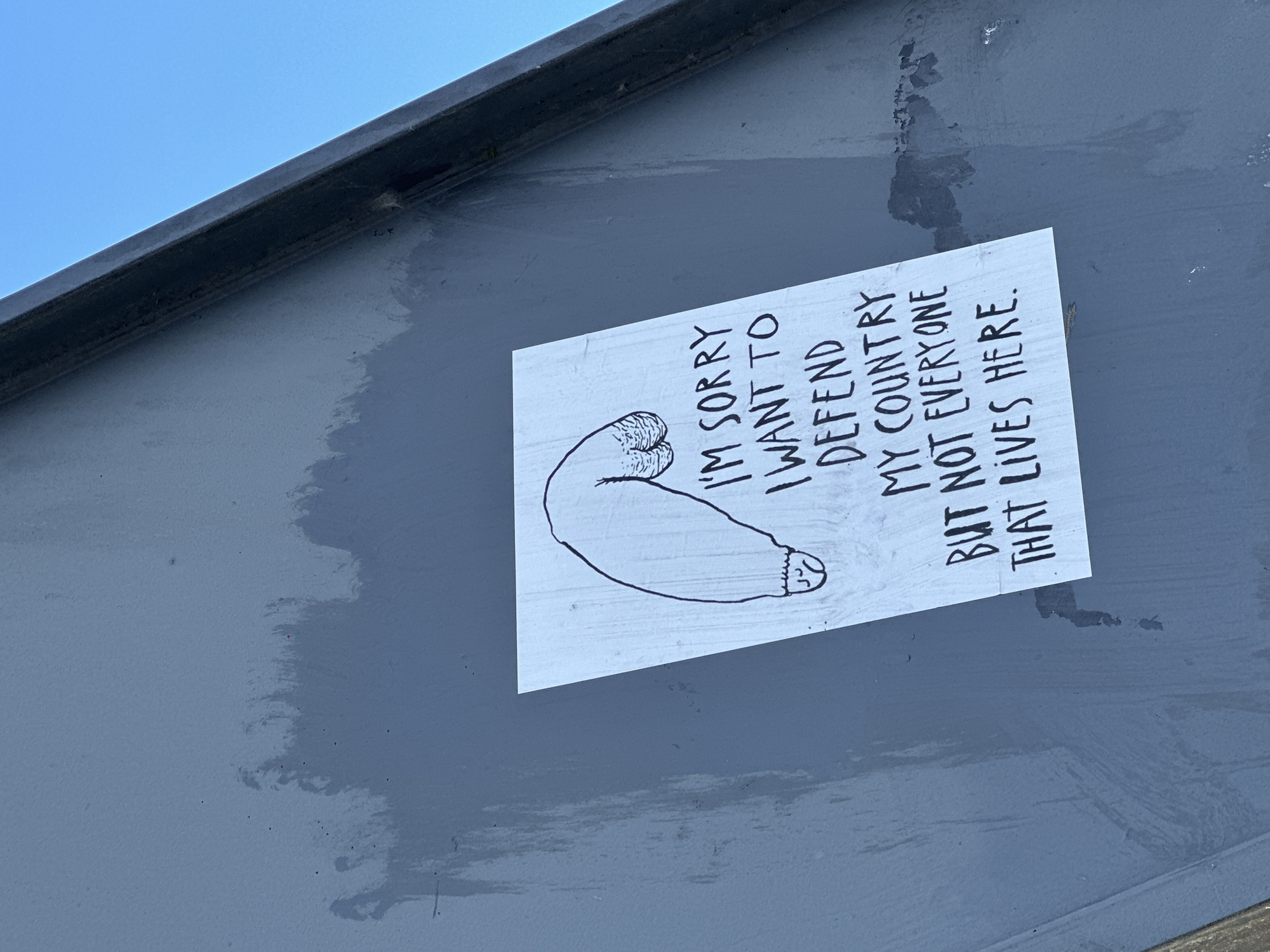 A sticker with a drawing of a penis and testicles is adhered to a gray, textured wall. Text on the sticker reads: "I'M SORRY I WANT TO DEFEND MY COUNTRY BUT NOT EVERYONE THAT LIVES HERE." The sticker is placed off-center in the lower right quadrant of the image. The background is a light blue sky. The wall shows signs of wear, with streaks of lighter gray and dark marks. The angle of the wall suggests it is part of an exterior structure, possibly a building or a large utility box. The image is taken outdoors during daylight. No specific location in Amsterdam, Netherlands, can be identified from the visual information provided.
