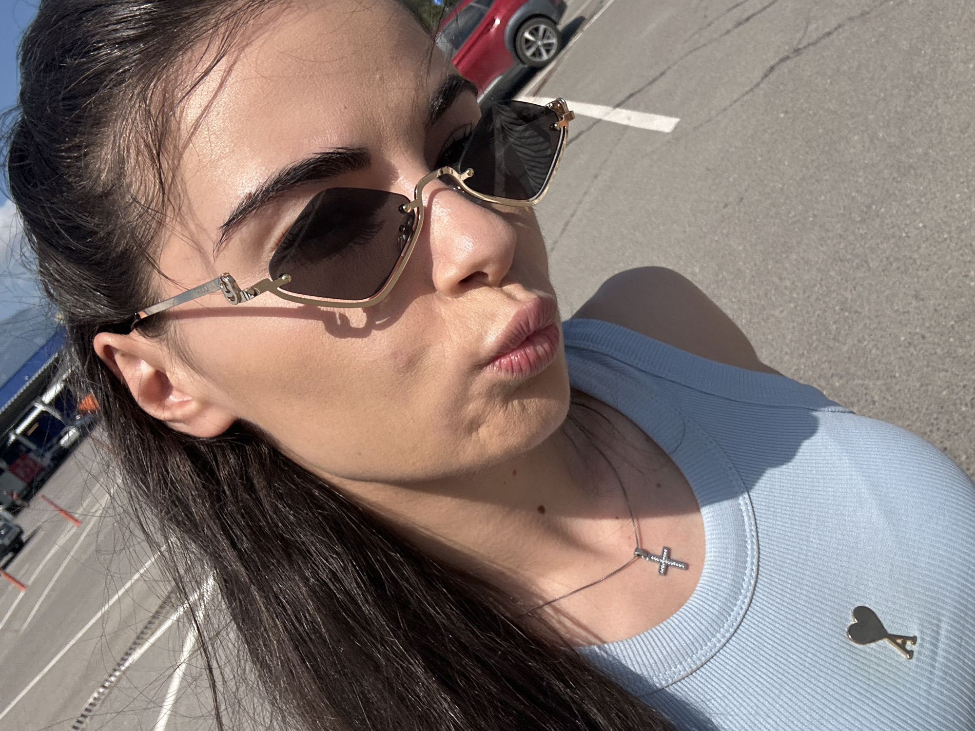 A close-up outdoor shot features a young female, situated in what appears to be a parking lot in Sofia, Bulgaria, under bright sunlight. Her long, dark brown hair flows over her right shoulder. She wears gold-framed Gucci sunglasses with dark, rectangular lenses, obscuring her eyes. Her lips are pursed, and she wears a light blue ribbed tank top. A thin silver chain necklace with a pave-set cross pendant rests on her chest, and a gold-colored Ami de Coeur heart-shaped pin is attached to the left side of her top. The background consists of grey asphalt with white parking lines. A red compact SUV is partially visible in the mid-ground right, with further indistinct vehicles and commercial buildings in the far distance. Strong shadows are cast on her face and upper body.