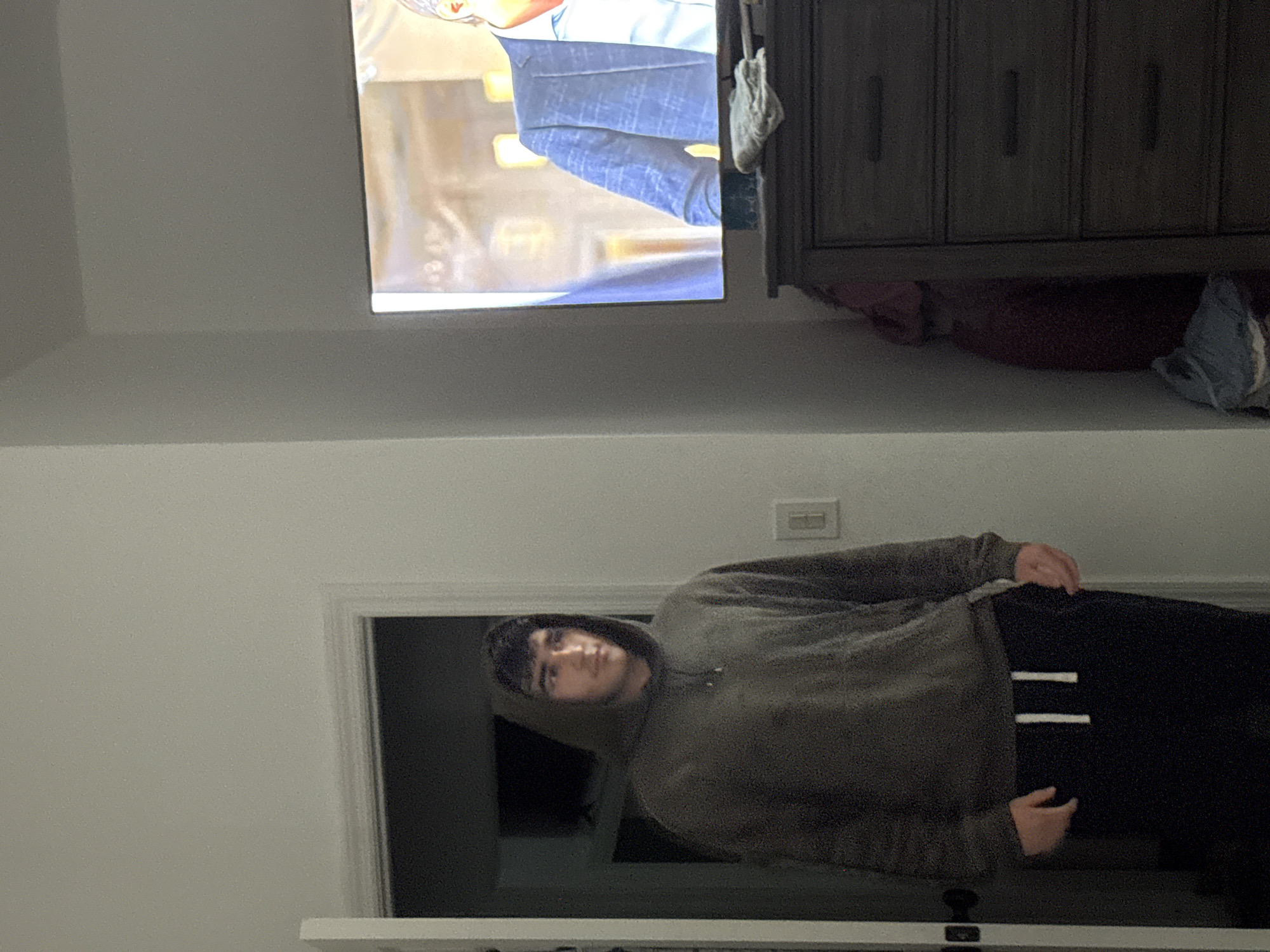 A young man stands in a doorway looking off to the side, wearing a dark green hooded sweatshirt and black sweatpants. A television is visible in the background, displaying a man in a blue checkered suit. To the right of the television is a dark dresser. The room appears to be a bedroom in a residential setting. The image was taken in Bellmore, United States of America.