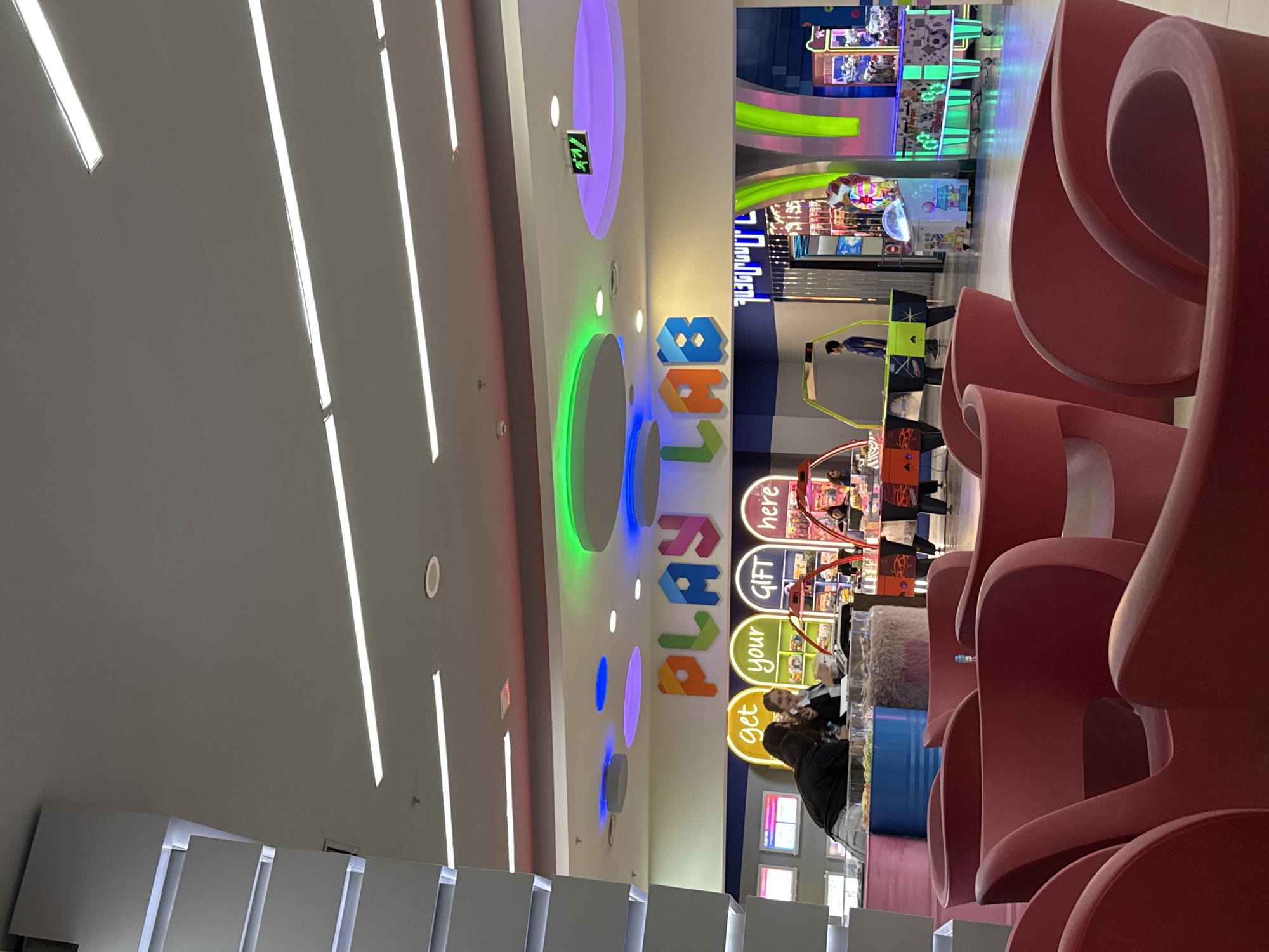 The image depicts an indoor entertainment or arcade area, likely within a shopping mall. The perspective is tilted, looking across and slightly upwards at the scene.
In the foreground and mid-ground on the right, a series of large, sculptural red seating elements with flowing, curved designs are prominent. To their left, a brightly lit counter or service desk is visible, where several people are gathered. One person in a dark jacket is seated facing the counter, while another individual with light hair is behind it, possibly an attendant.
The background features a vibrant arcade with numerous game machines. A person in a blue shirt is standing by an air hockey table. Other arcade games with flashing lights and various prizes are visible further back.
The ceiling above the counter area is white, adorned with modern lighting fixtures, including long linear lights and circular lights that cast colorful glows in green, blue, and purple. A large, colorful sign spells out "Play Labs" on the wall, with smaller, indistinct text below it. Adjacent to "Play Labs," three circular signs are stacked vertically, reading "get," "your," and "GIFT here." A green exit sign with an arrow and Arabic/Farsi text "مخرج" (meaning "exit") is also visible on the ceiling.
The overall atmosphere is bright and inviting, typical of a modern entertainment venue. The time of day is not discernible as the space is artificially lit. The setting suggests an active leisure environment.