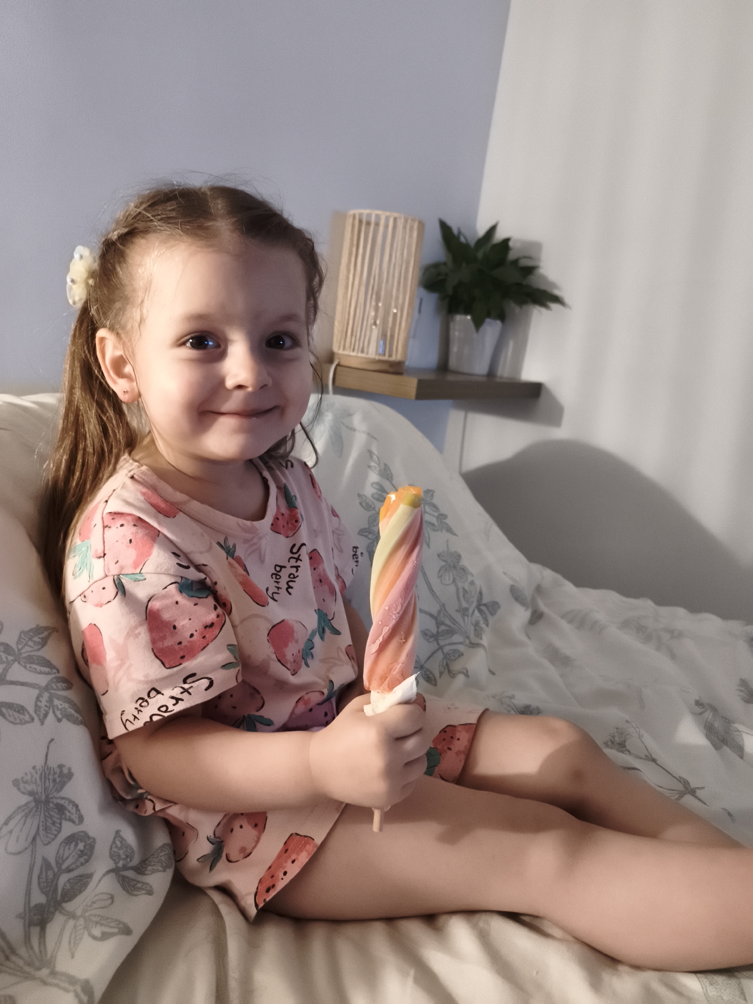 A young girl with brown hair in a ponytail, adorned with a white hair clip, sits on a bed. She is wearing a pink t-shirt and shorts, both featuring a strawberry pattern. In her right hand, she holds a colorful, swirled popsicle. Her expression is a cheerful smile, and she is looking directly at the camera. The background shows a wall with a shelf holding a lamp and a potted plant. The bed is covered with white bedding that has a subtle floral pattern. The lighting suggests it could be evening or indoors with artificial light. The overall mood is relaxed and happy. The word "strawberry" is visible in text on the girl's t-shirt.