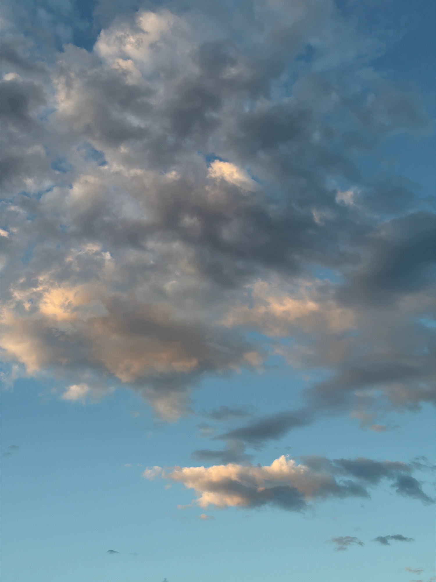This is a vertical, eye-level, full shot of a cloudy sky. The clouds are dark gray and white, with hints of pink and orange from the setting sun. The sky is a light blue color. There are no people, subjects, or objects in the image, and no actions or interactions are taking place. The scene is purely natural and appears to be an outdoor environment.