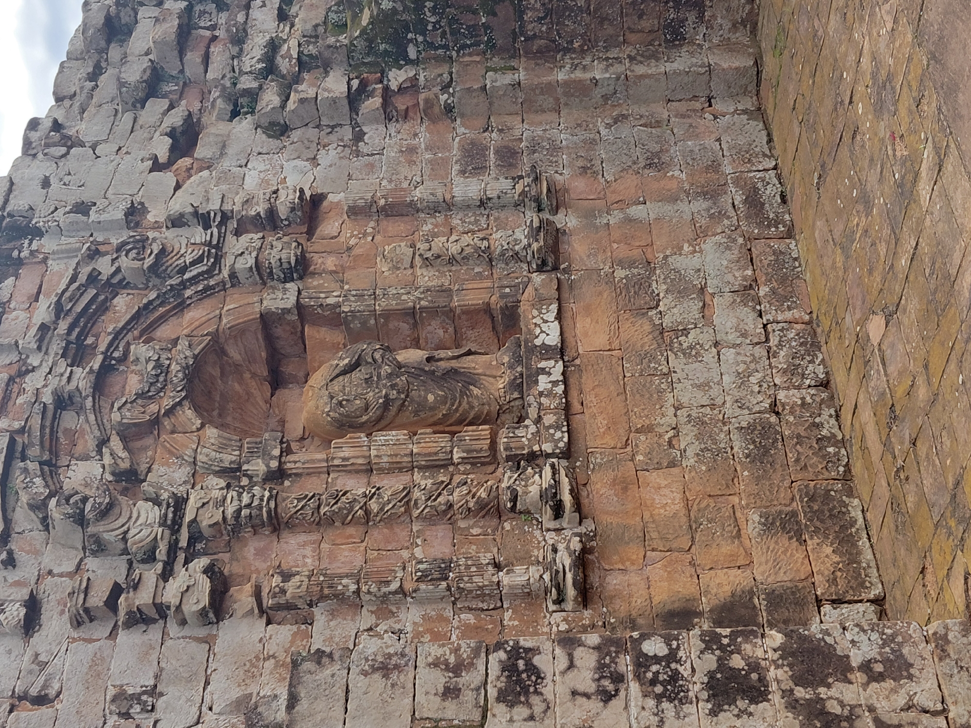 The image depicts the ruins of an ancient stone structure, likely a religious building, with a weathered and aged appearance. The masonry is constructed from large, roughly hewn stone blocks, predominantly in earthy tones of brown, orange, and gray. Some blocks are covered in patches of white and dark lichen or moss, indicating exposure to the elements over a significant period.
The central feature is a recessed alcove or niche within the wall, housing a headless stone statue of a figure draped in robes. The statue is positioned on a pedestal and flanked by decorative columns or pilasters carved with intricate patterns. Above the alcove, there is a semicircular arch with carved ornamentation.
The surrounding wall exhibits similar construction, with visible signs of erosion and decay. The texture of the stone is varied, with some surfaces appearing smooth while others are rough and pitted. In places, mortar is visible between the stones.
The foreground shows a paved floor made of rectangular stone tiles. These tiles are also aged and weathered, with some discoloration and minor damage. The overall environment suggests an outdoor historical site, likely in a climate that supports moss and lichen growth.
This structure is located in the Jesuit Missions of La Santísima Trinidad de Paraná, a UNESCO World Heritage site near the city of Trinidad, Paraguay. This particular architectural detail is part of the ruins of the historic church within the mission complex.