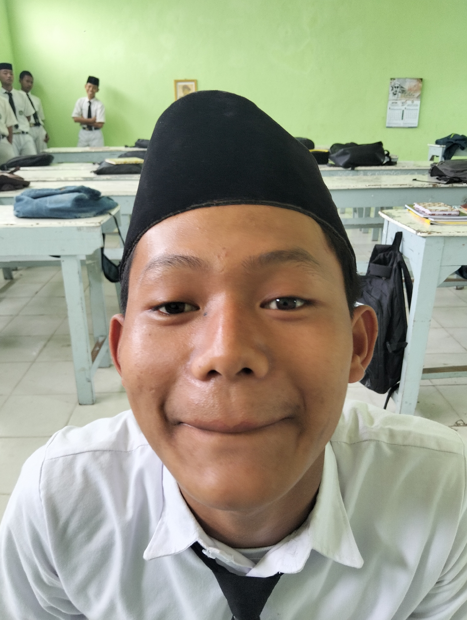 The image captures a young male student wearing a white collared shirt, a black tie, and a black peci (a type of cap) in what appears to be a classroom. He is positioned centrally and is looking directly at the camera with a subtle smile. In the background, other students are visible, some in similar attire. Desks are arranged in rows, and a calendar hangs on a light green wall. The overall setting suggests an educational environment, possibly in Kranji, Indonesia, given the context provided. The lighting is bright, and the focus is primarily on the student in the foreground.