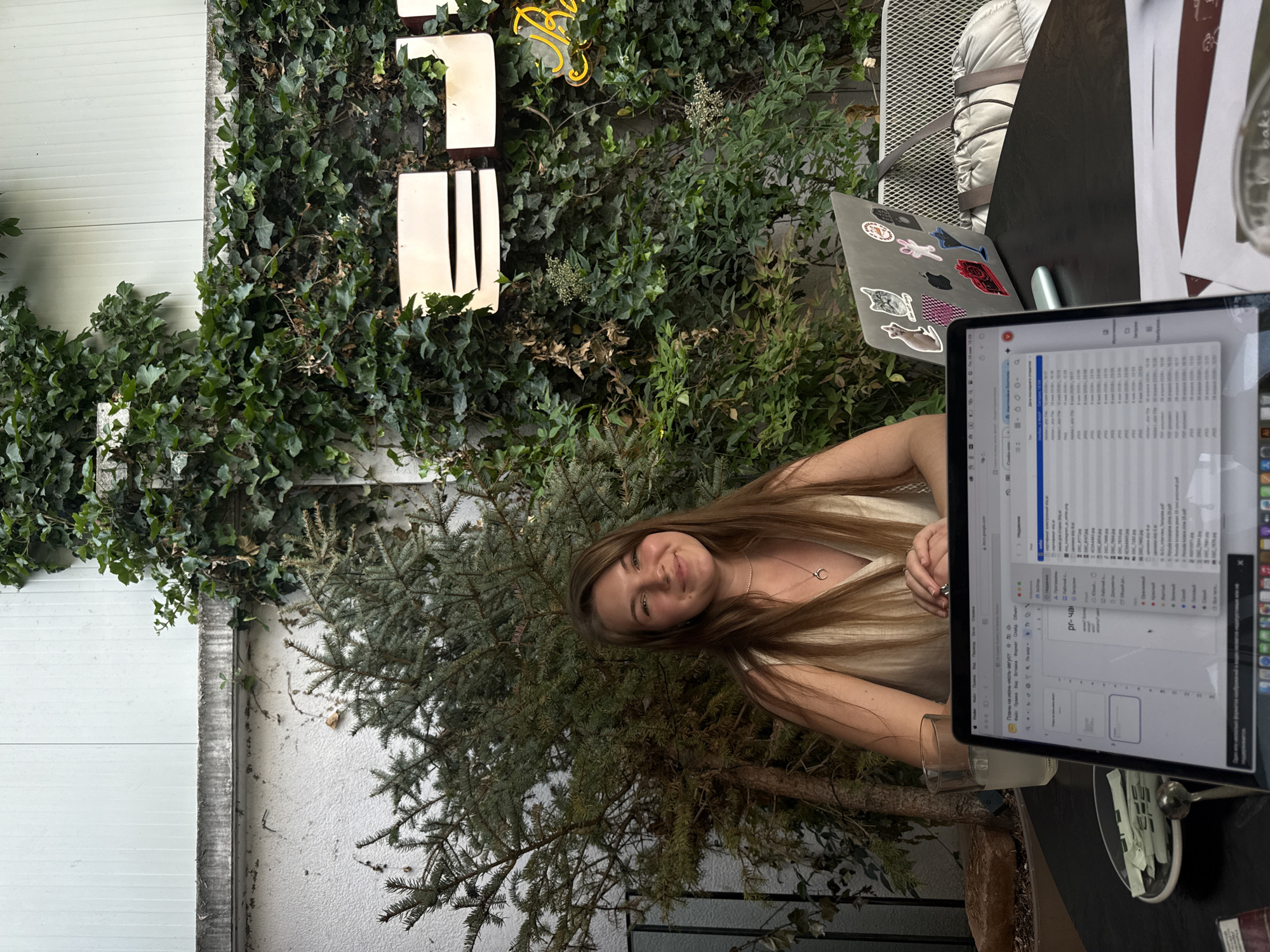 The image shows a young woman sitting at an outdoor table, likely a café or restaurant, in Belgrade, Serbia. She is working on a laptop with documents open on the screen. The laptop has stickers on the back. She is sitting amongst greenery, including ivy on a wall and a small pine tree. A white wall can be seen on the left. A metal sign is attached to the green wall.
