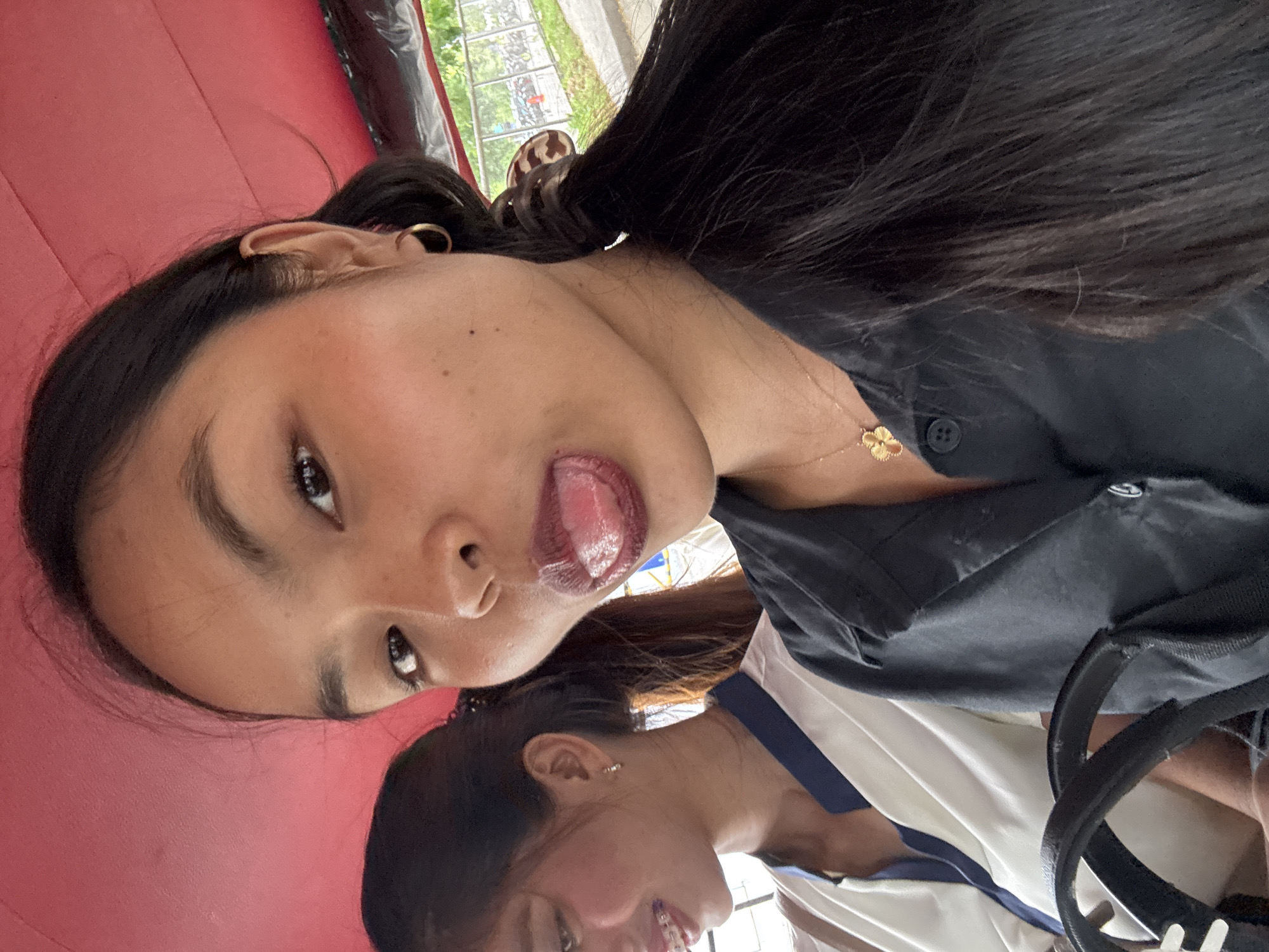The image captures a selfie of two young women in what appears to be the interior of a vehicle, likely a tricycle given the red roof visible. The woman in the foreground, facing the camera, is sticking her tongue out and wearing a black collared shirt with a golden clover pendant necklace. Her hair is dark and pulled back. To her left, another young woman is visible, looking away from the camera and smiling. She is wearing a white shirt with a dark blue collar and has braces on her teeth. The background, seen through a window or open side of the vehicle, shows a street scene with some greenery and parked vehicles, suggesting an outdoor urban environment. This scene is consistent with the common modes of transportation and street views found in Davao City, Philippines.