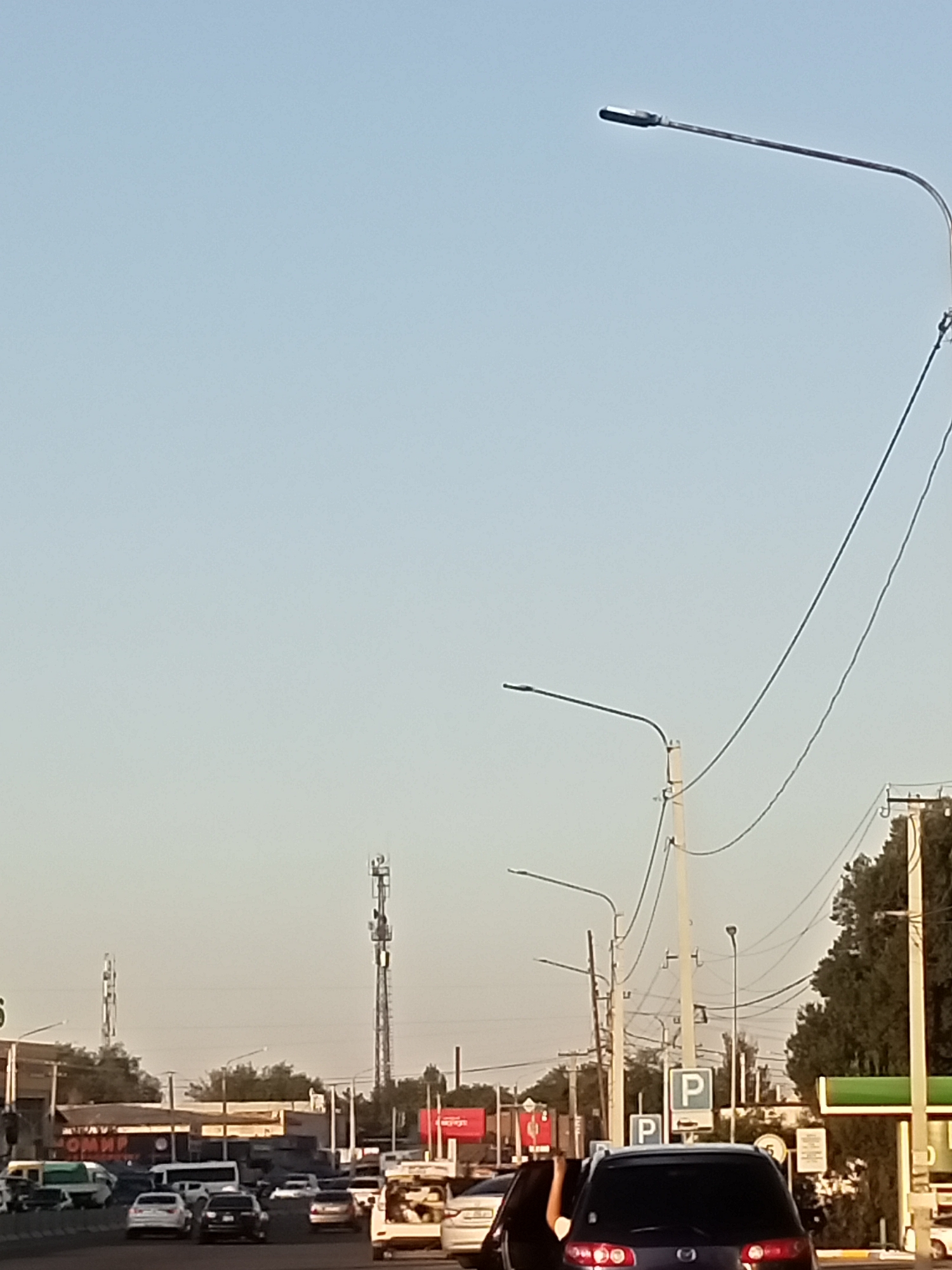 The image captures a daytime scene on a city street in Sokuluk, Kyrgyzstan, with a clear sky dominating the upper portion of the frame. The lower half of the image depicts a busy road with several vehicles. A light blue sedan is visible in the foreground, to the left, followed by a dark SUV. Further down the road, a line of cars including a white sedan and a dark SUV are stopped in traffic. On the right side of the image, a white minivan with its rear door open is parked.
Several streetlights on poles are present, some with visible wires. In the background, two tall communication towers rise above the buildings. The buildings themselves appear to be commercial establishments, with some signage visible, including what appears to be "OMMR" in Cyrillic on a building to the left. There are also trees and utility poles lining the street. Two parking signs with the letter "P" are visible. The overall impression is of a typical urban environment during daylight hours.