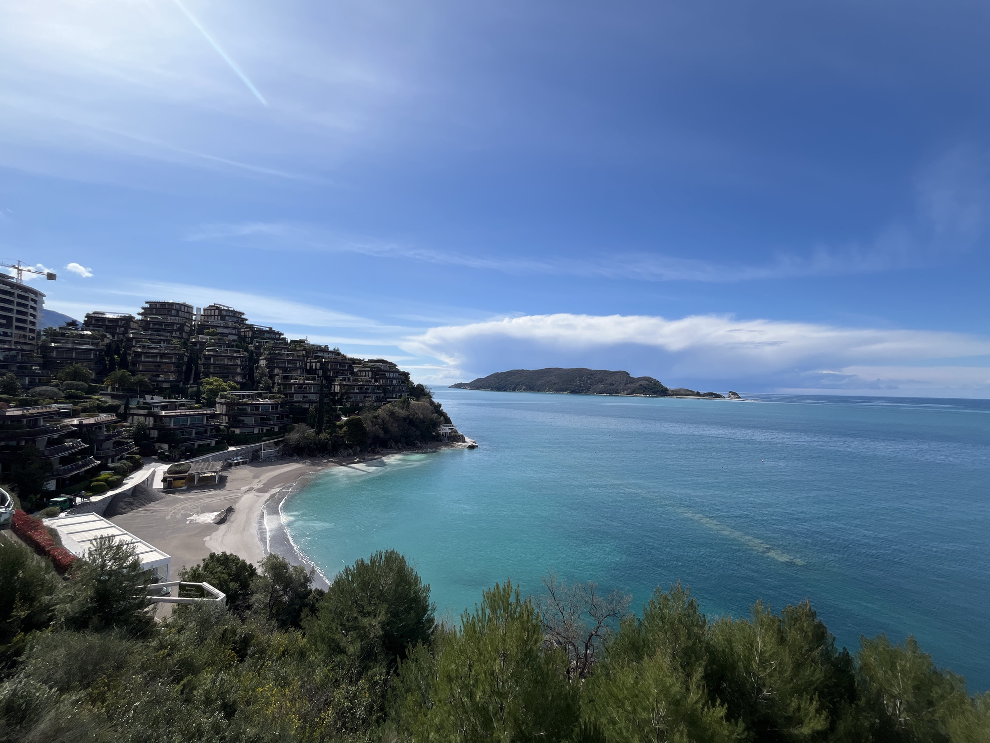 This wide-angle coastal view captures the area of Dukley Gardens and Caprice Beach in Budva, Montenegro, looking eastward towards Sveti Nikola Island under a bright blue sky with scattered thin white clouds. A prominent sunbeam extends diagonally from the upper left.
In the foreground, dense green foliage, primarily shrubs and coniferous trees, obscures the lower portion of the image, indicating a high vantage point.
The midground features a gently curving, light-colored sand and pebble beach, where small white waves break against the shore. Directly behind the beach are several modern, multi-story terraced buildings with dark frames and extensive glass windows, integrated into the steep green hillside. A winding path or road runs along the base of these structures. A white, geometric building with a walkway is positioned closer to the beach, and a smaller yellow structure is visible nearby. In the far upper left, a partially constructed high-rise building with exposed concrete is visible, topped by a yellow tower crane.
The vibrant turquoise waters of the Adriatic Sea dominate the center, transitioning to a deeper blue further from shore. A subtle, elongated linear pattern, possibly an underwater structure or sandbar, is visible beneath the surface in the deeper water. A small, reddish buoy floats in the mid-distance.
In the background, a large, dark green, undulating island, Sveti Nikola Island, stretches across the horizon, separating the sea from the distant sky. No people or active interactions are visible within the scene.