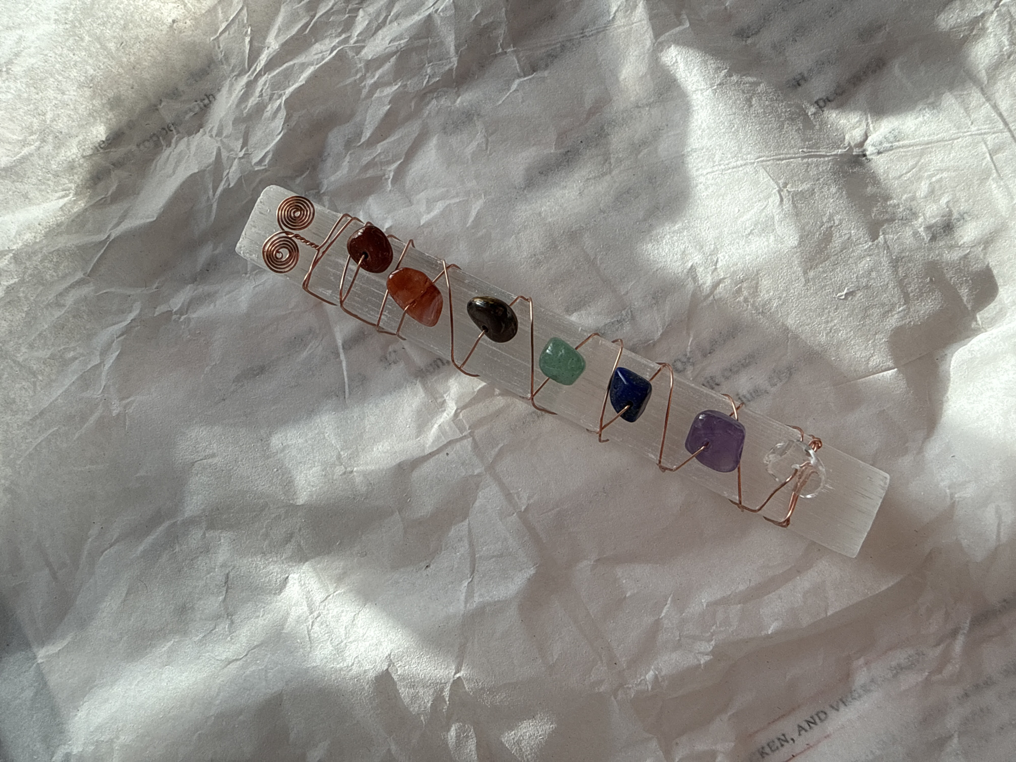The primary subject is a clear, rectangular mineral stick, resembling selenite, adorned with seven small, tumbled, colorful stones arranged in a linear sequence. These stones, from left to right, appear to be reddish-brown, orange, dark brown/black, green, blue, purple, and clear. They are secured to the mineral stick by intricate copper wire wrapping, which includes decorative spirals at the left end of the stick.
There is no specific activity or event taking place; the image is a still-life photograph of the crafted object.
The setting consists of a crinkled, light-colored paper background, likely white or off-white tissue paper, which displays various folds and textures. Bright, natural lighting, possibly sunlight, illuminates the scene, casting distinct highlights on the paper's creases and creating subtle shadows. This strong illumination suggests the photograph was taken during the daytime. The overall presentation implies a handcrafted or spiritual item. There are no specific visual cues to indicate the location as Solana Beach.
Faint, partial, and largely illegible text is visible embedded within the crinkled background paper, appearing as watermarks or printed matter. Fragments such as "REN, AND VEG", "with", "Dot", and "Dec" can be discerned, but the full context is obscured.