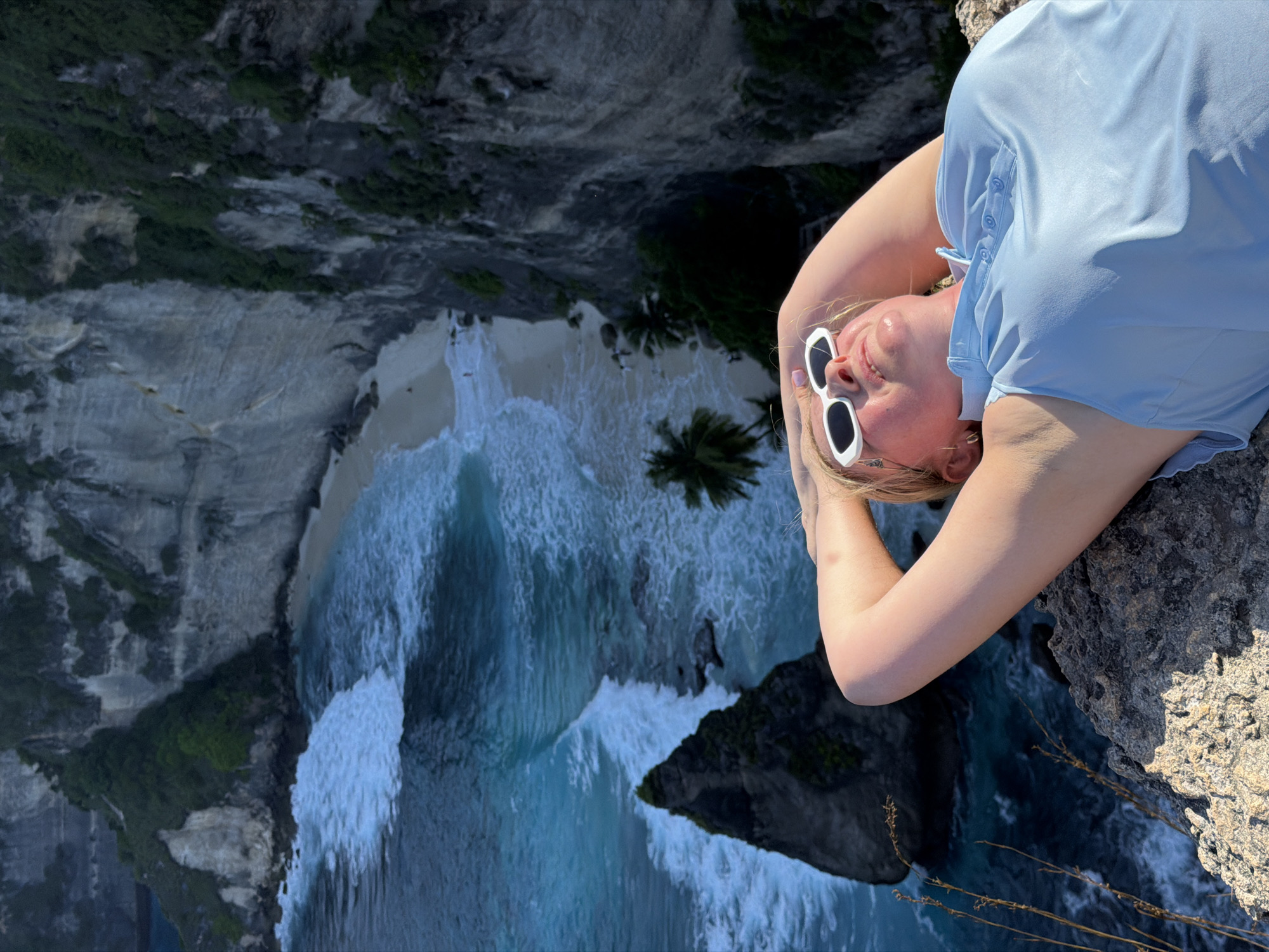 A young woman with blonde hair and white-rimmed sunglasses lies on her back on a rocky cliff edge, with her hands behind her head. She wears a light blue polo shirt. The background depicts a dramatic coastal landscape in Nusa Penida, Indonesia. She is overlooking a beach with turquoise water and crashing waves against a jagged rock formation in the sea. Steep, verdant cliffs rise behind the beach, with scattered palm trees visible. The scene is bathed in bright sunlight.