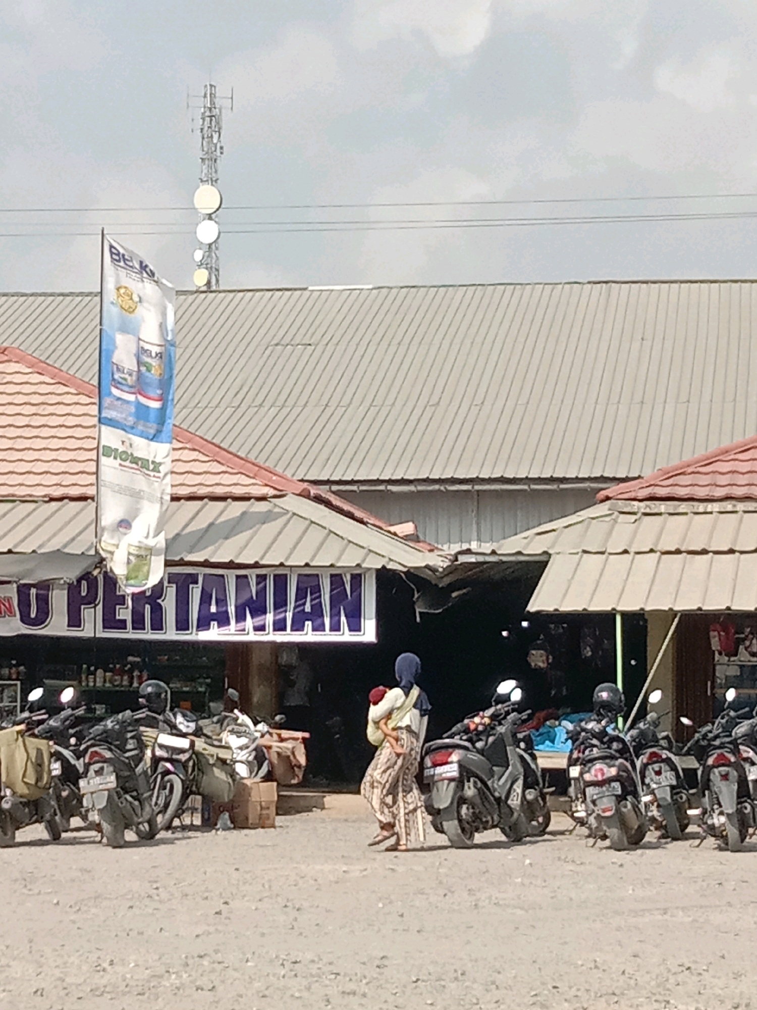 The image captures an outdoor scene in Fajar Mataram, Indonesia, likely near an agricultural supply store. In the background, a cell tower stands against a cloudy sky. The main subject is a row of parked motorcycles, some equipped with saddlebags. A woman wearing a headscarf and a patterned skirt is visible in the mid-ground, carrying a baby on her back. She appears to be walking away from the motorcycles and towards a building with a red-tiled roof and a sign that reads "U PERTANIAN" (Agriculture). A banner with product images and text is hanging from the building's overhang. The ground is covered in gravel. The overall impression is that of a bustling local market or commercial area catering to agricultural needs.