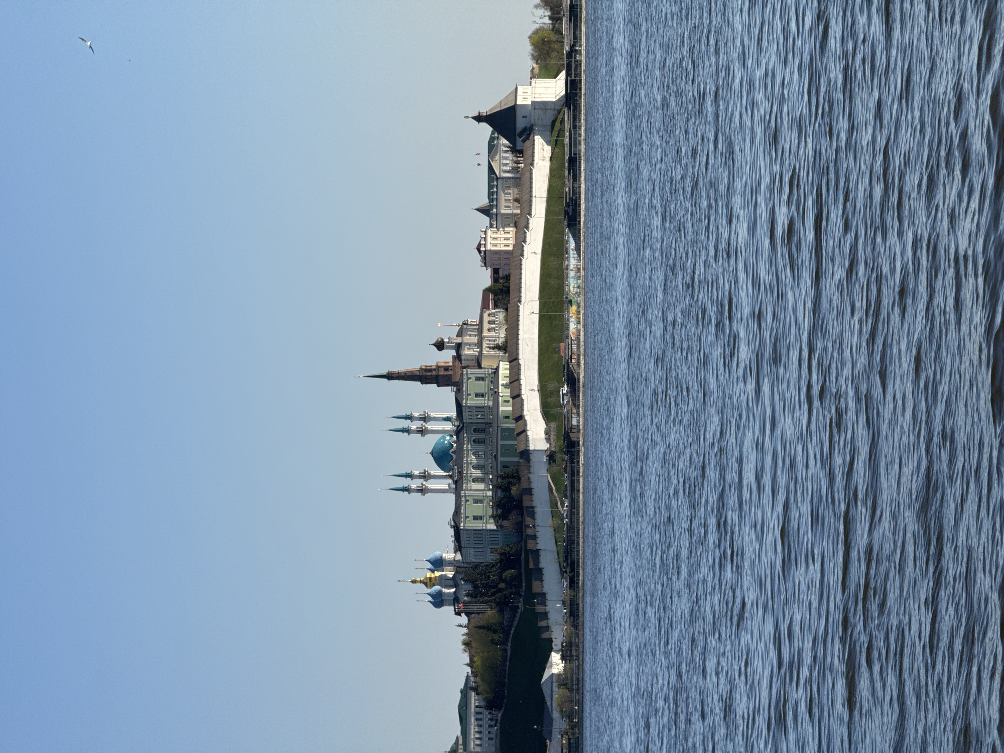 The image shows a cityscape along a body of water, likely taken in Kazan, Russia. Notable architectural features include buildings with a mix of colored domes and spires, and a white wall extending along the waterfront. The sky is a clear, light blue, and a single bird is visible in the upper left corner. The water has a rippled surface texture.